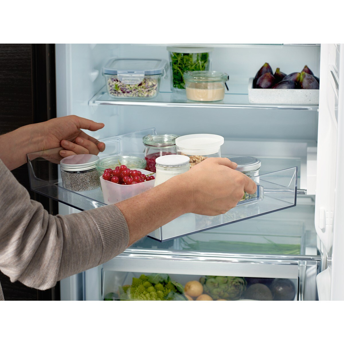 Osoba trzymająca półkę lodówki ze słoikami z jedzeniem. Inne półki pokazują figi, jagody i warzywa.