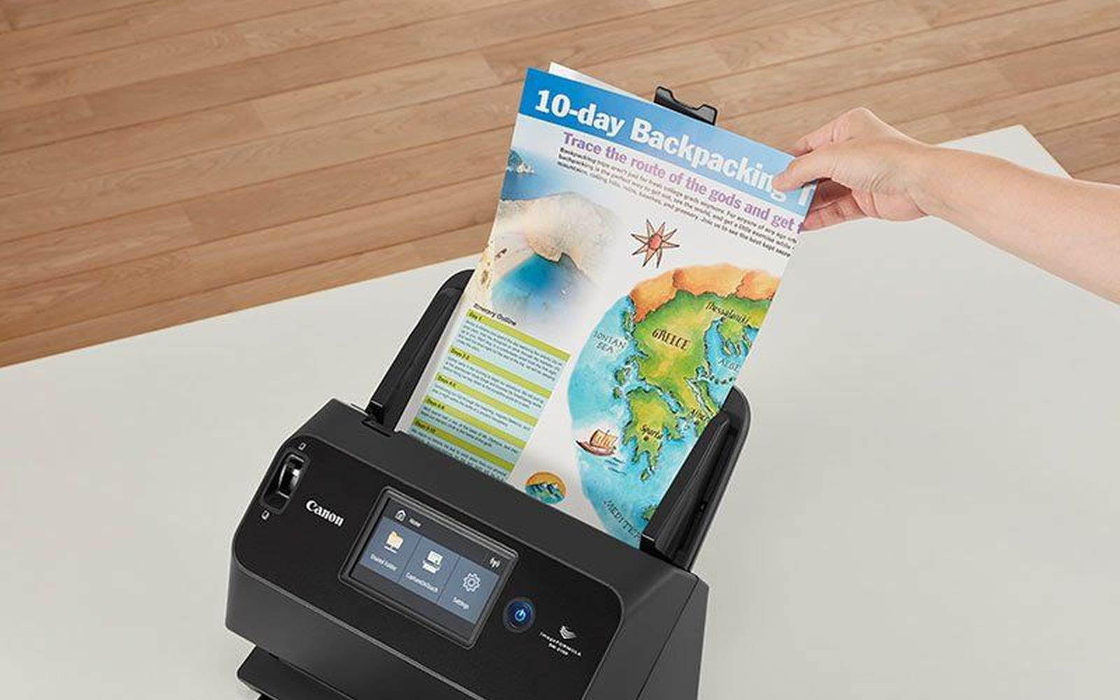 Ręka wkłada kartkę papieru do skanera Canon. Papier to 10-dniowy przewodnik po backpackingu z mapą Grecji.