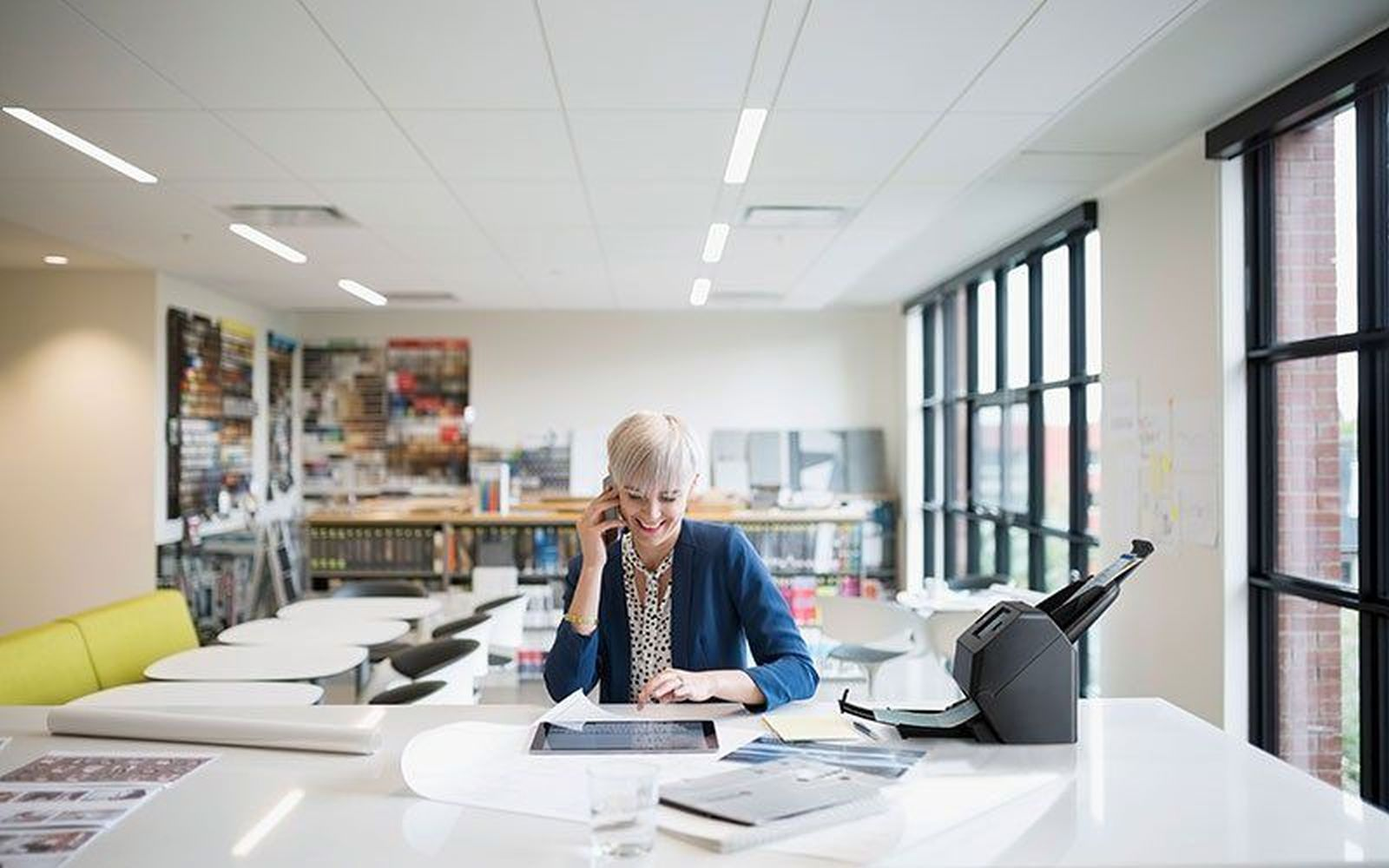 Kobieta w niebieskiej kurtce rozmawia przez telefon, pracując na tablecie przy białym biurku. Okna i półki w tle.