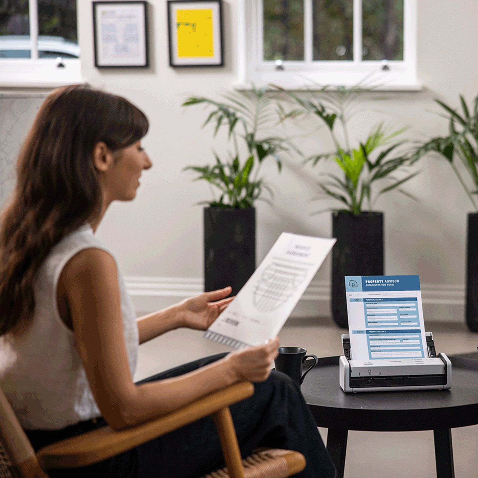 Kobieta siedzi, trzyma papiery, w pobliżu skanera i formularza. Rośliny w tle. Kubek kawy na stole.