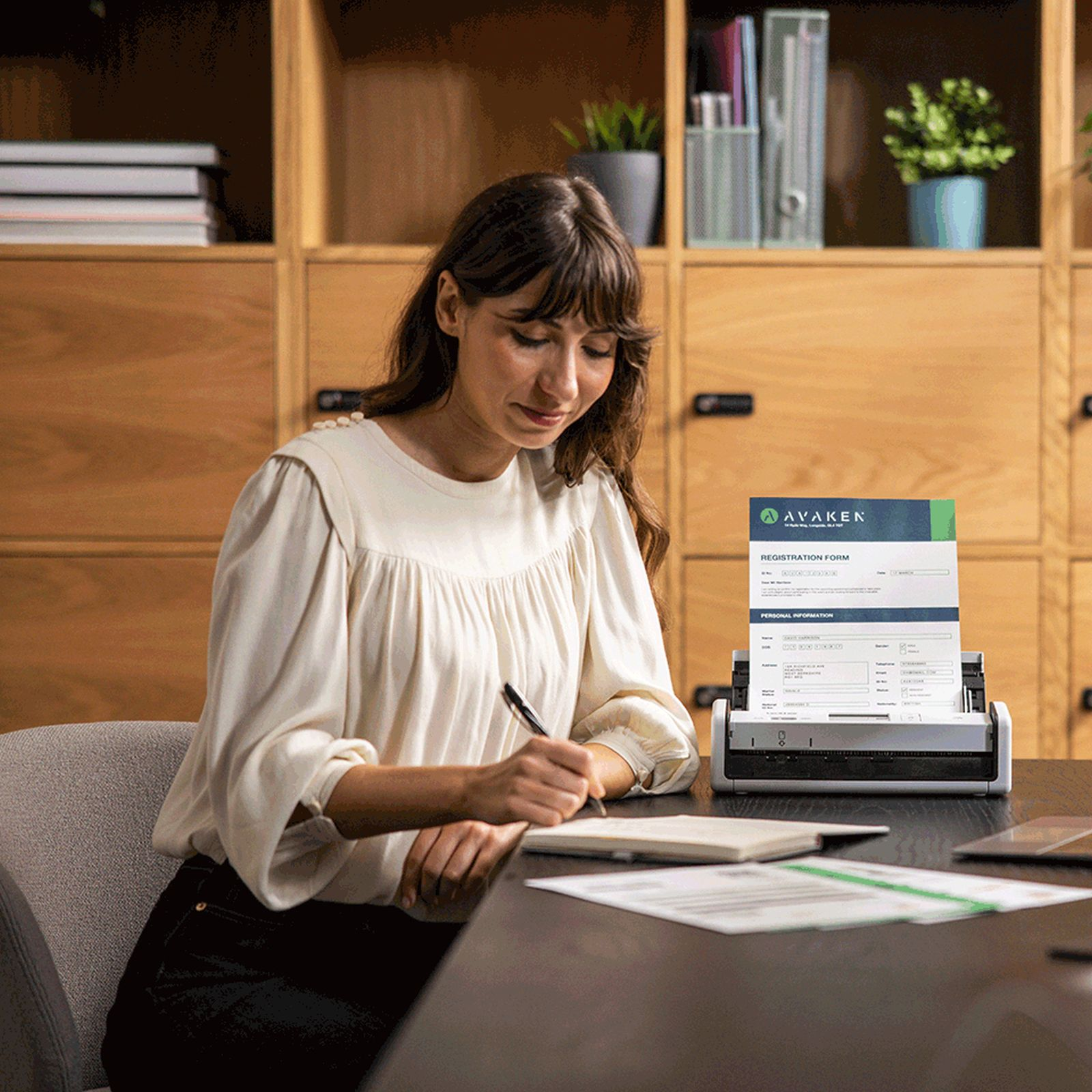 Kobieta pisze w notatniku, dokument w skanerze. Drewniane szafki w tle z roślinami.