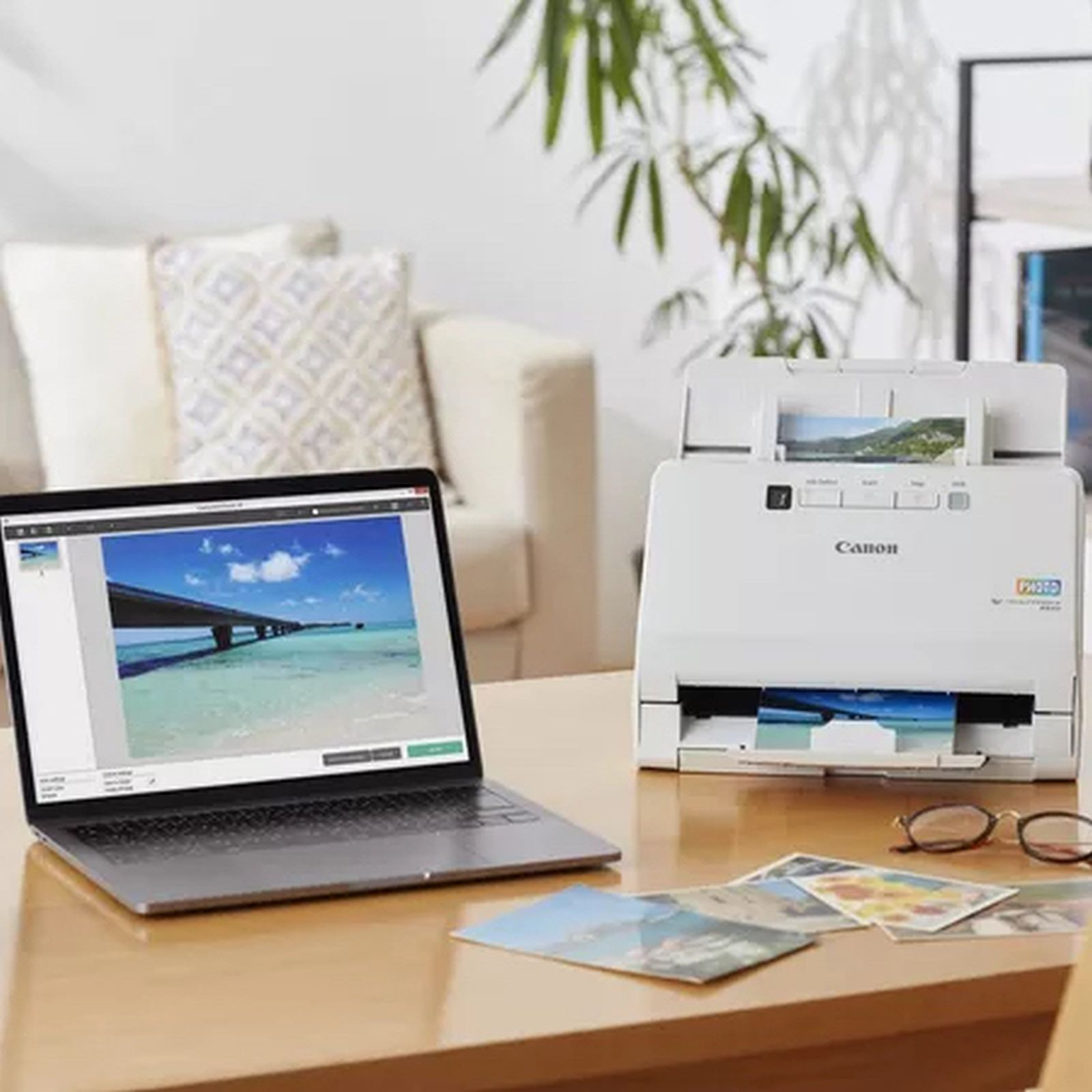 Laptop wyświetla obraz mostu nad wodą. Na stole znajduje się drukarka i zdjęcia. W tle jest sofa.