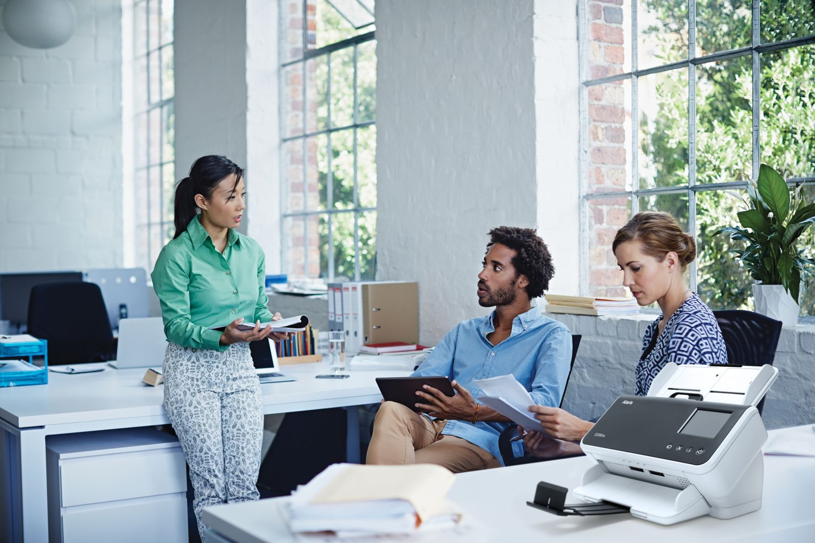 Trzech pracowników biurowych w jasnym biurze. Jedna kobieta mówi, trzymając notatnik. Dwóch innych słucha, jeden trzyma tablet.