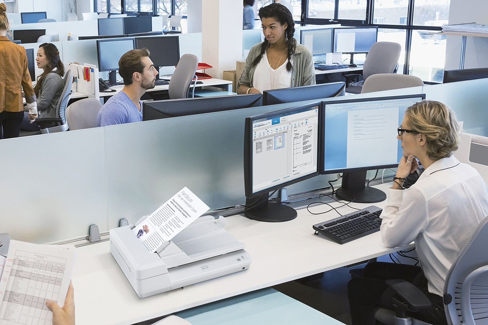 Scena biurowa. Ludzie przy biurkach, niektórzy patrzą na ekrany komputerów. Dokument jest skanowany. Biały skaner i ekrany.