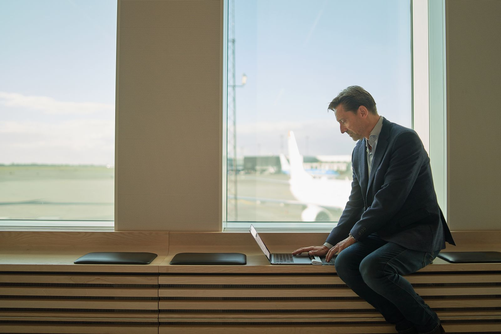 Man in a suit using a laptop near a window. An airplane is visible through the window. Wooden bench and cushions are in the front.