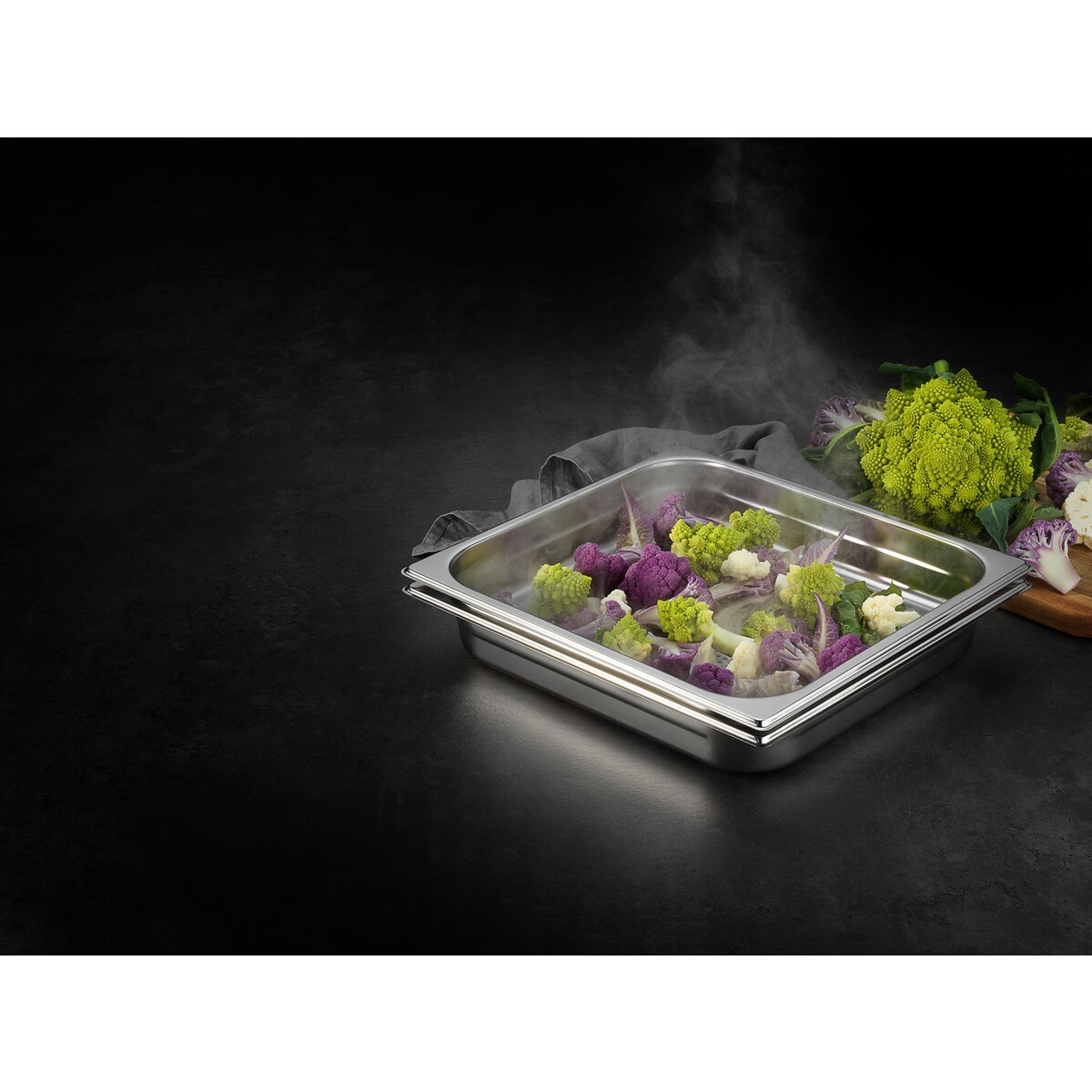 Dampfendes Gemüse in einer Metallschale. Lila, grüner und weißer Blumenkohl. Dampf steigt auf. Dunkler Hintergrund mit Holzbrett und mehr Gemüse.