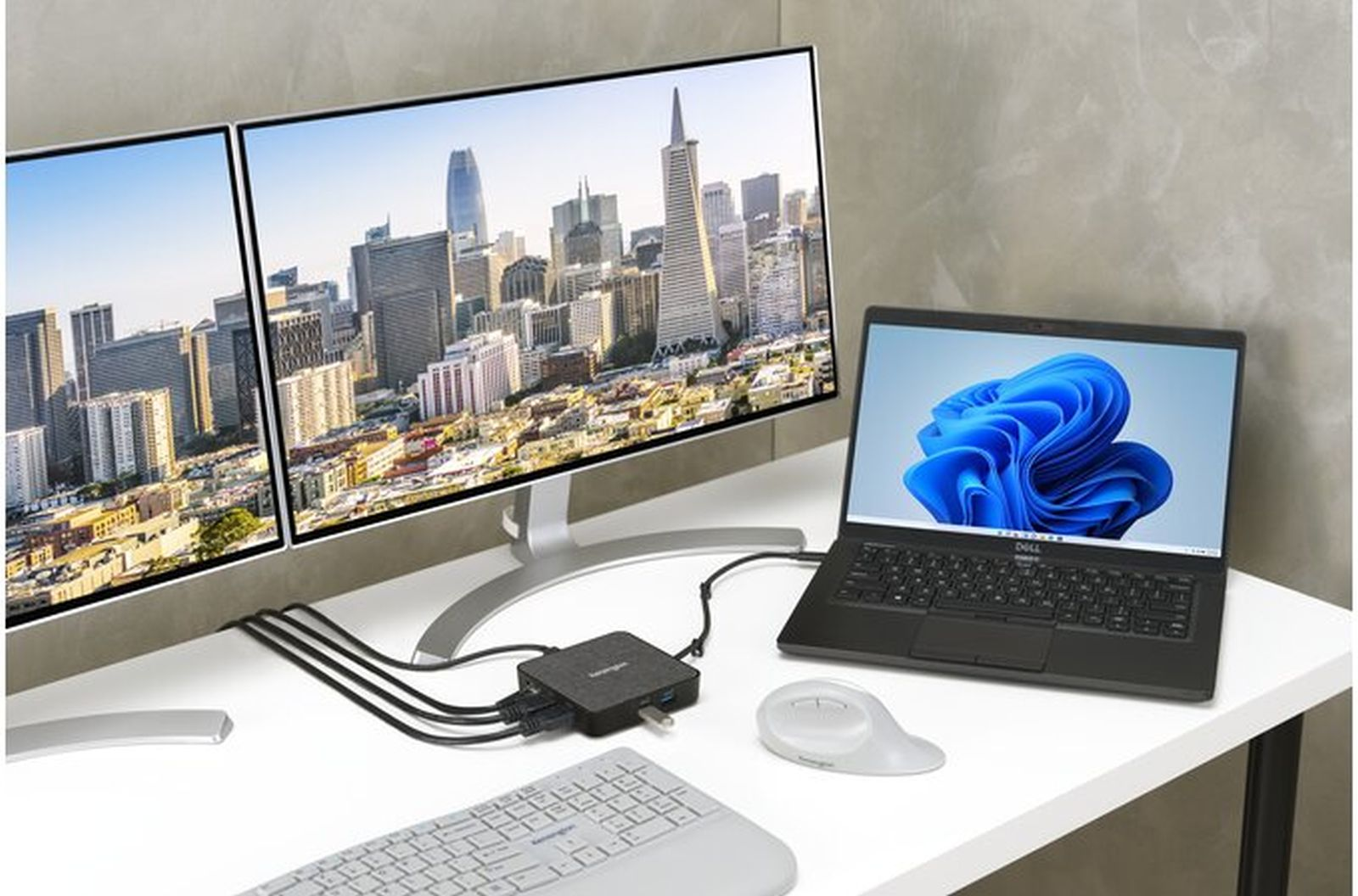 Ein Schreibtisch mit zwei Monitoren, die eine Stadtansicht zeigen, einem Laptop mit blauem Design und einer Tastatur.
