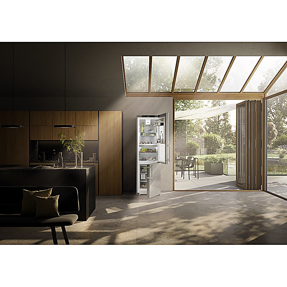 Cocina moderna con nevera abierta. Ventanas grandes muestran un patio. Armarios oscuros y una isla negra.