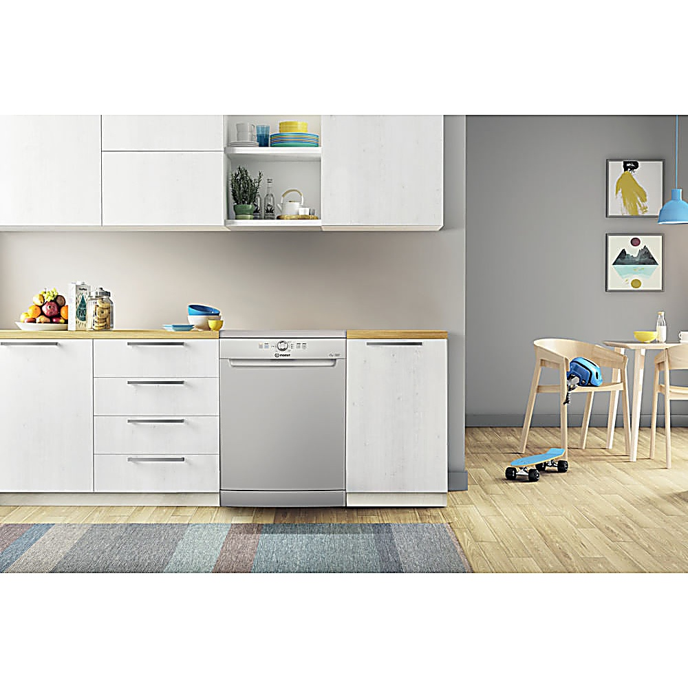 Cucina con mobili bianchi, lavastoviglie argentata, tavolo in legno con sedie, skateboard e tappeto colorato sul pavimento.