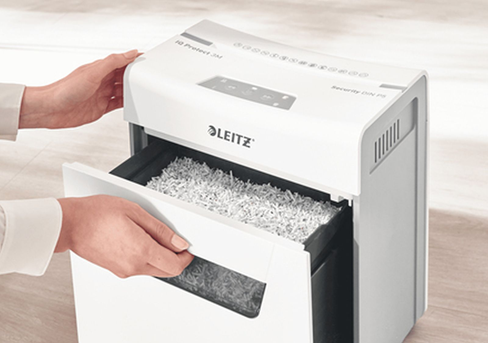 Osoba otwiera białą szufladę niszczarki papieru wypełnioną pociętym papierem. Niszczarka posiada logo Leitz.