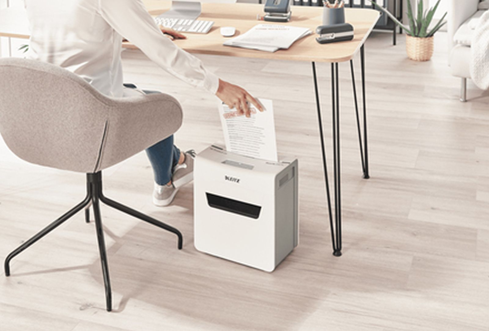 Osoba niszczy dokument. Biała niszczarka do papieru stoi na podłodze obok biurka. Osoba siedzi na szarym krześle.