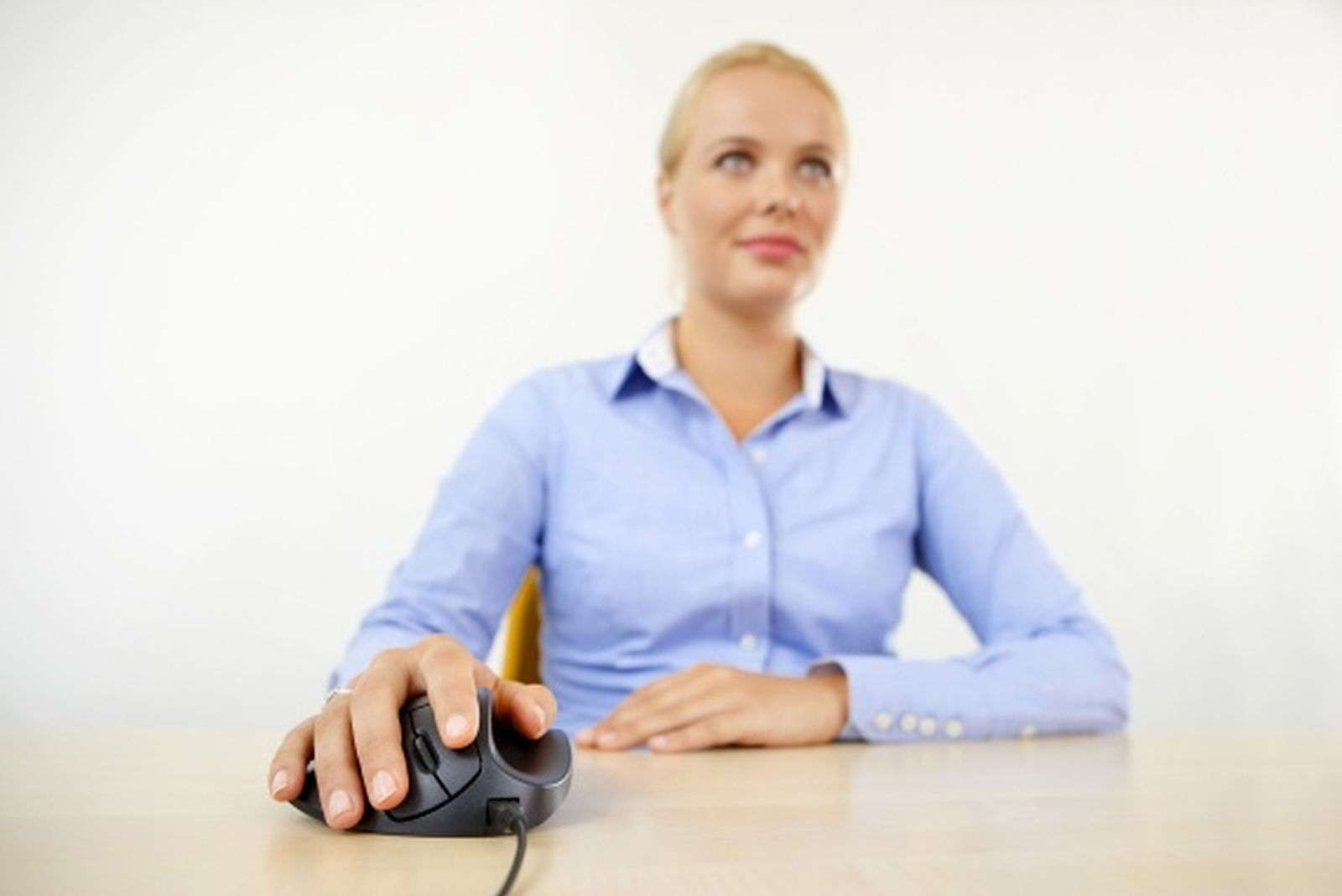 Kobieta w niebieskiej koszuli używa czarnej myszy na biurku. Tło to gładka biała ściana.
