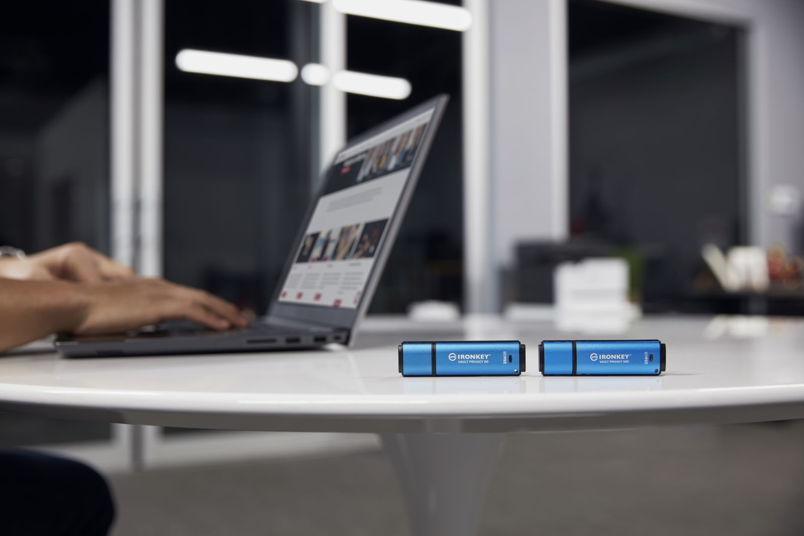 Osoba pisze na laptopie. Dwa niebieskie dyski USB Ironkey na białym stole. Nowoczesna przestrzeń biurowa w tle.