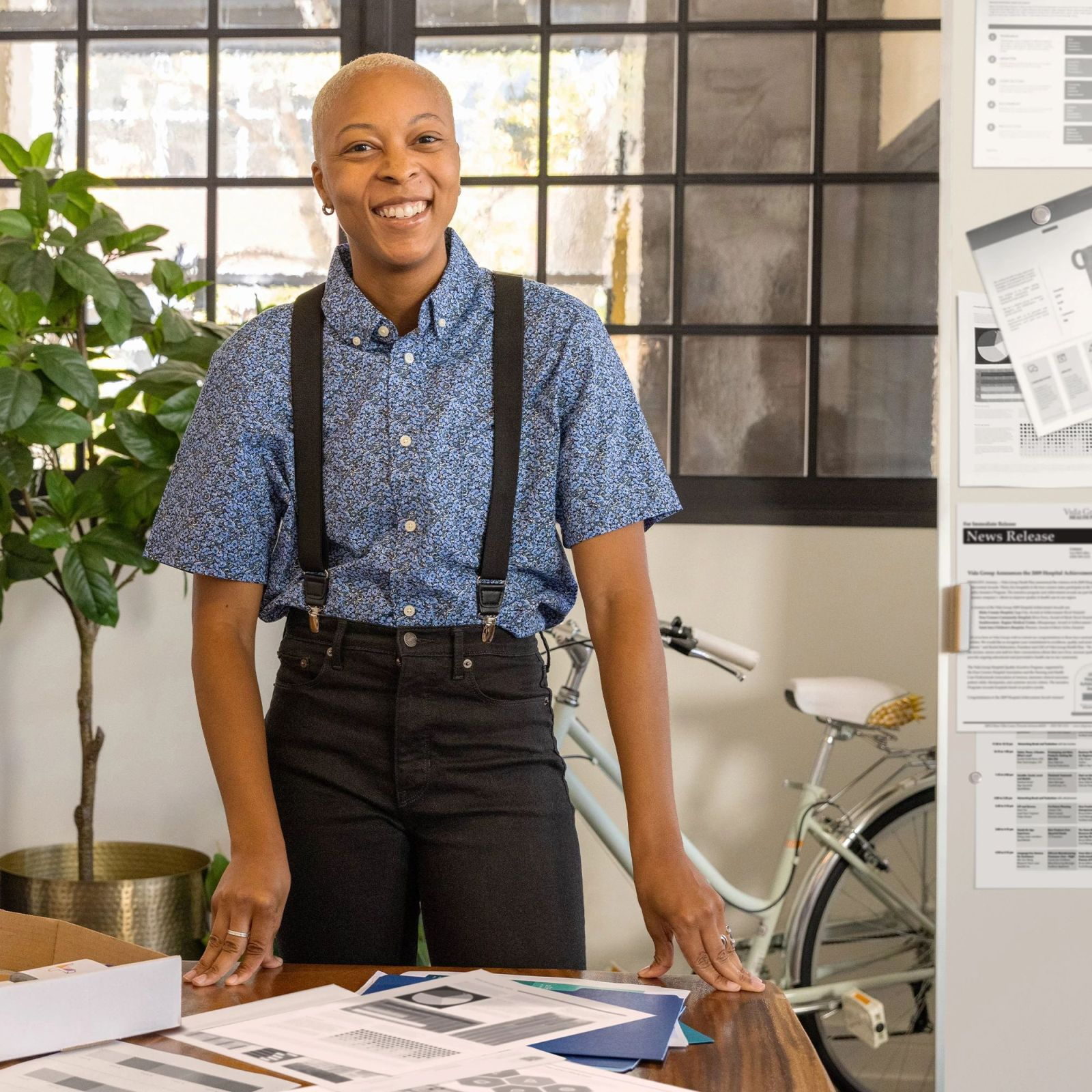 Osoba uśmiecha się, ubrana w koszulę we wzory i szelki, stojąca przy stole z papierami, rośliną i rowerem.