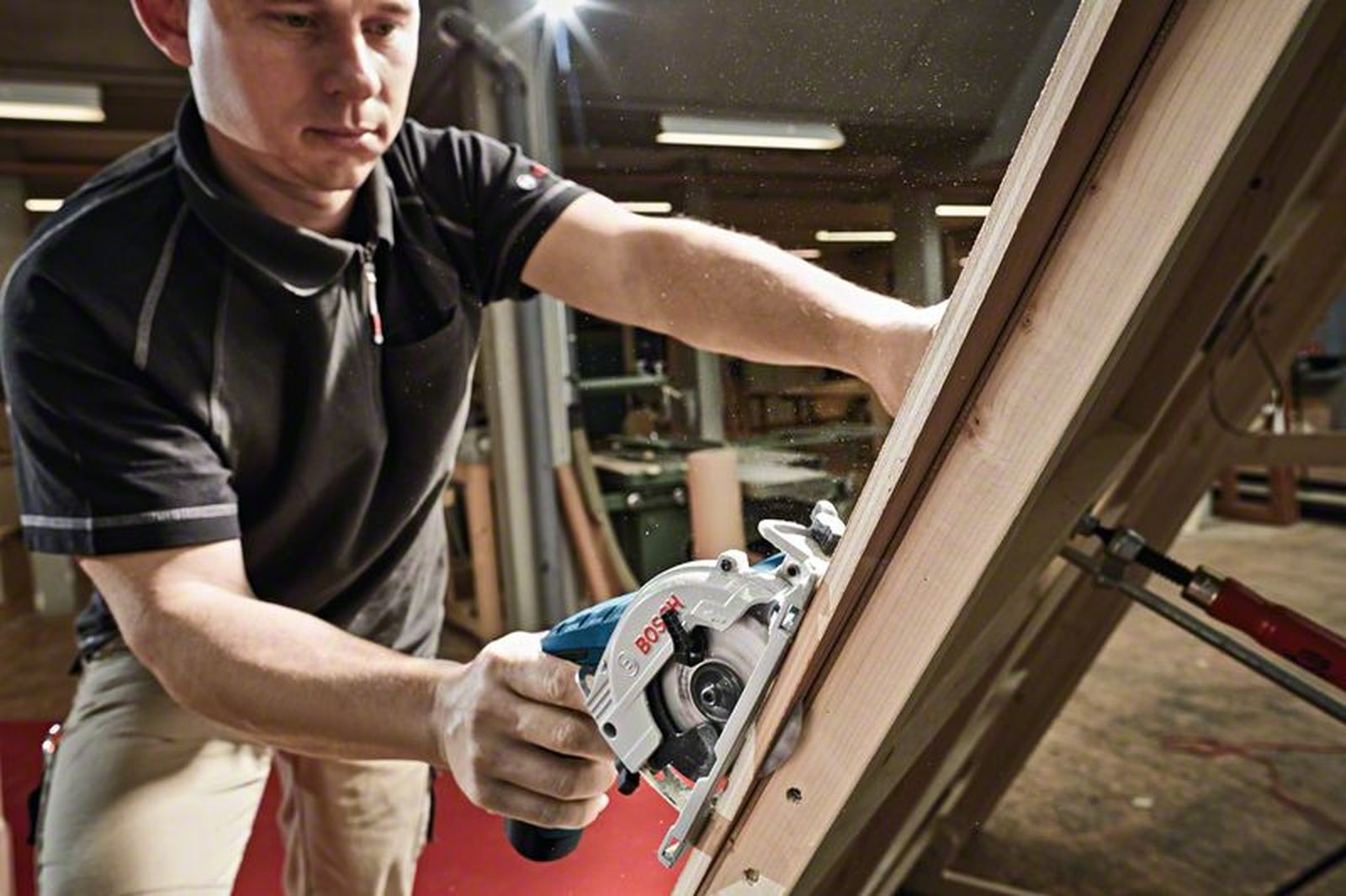 Mężczyzna używa piły tarczowej do cięcia drewna. Piła jest biało-niebieska. Pył leci. Mężczyzna ma na sobie czarną koszulę i beżowe spodnie.