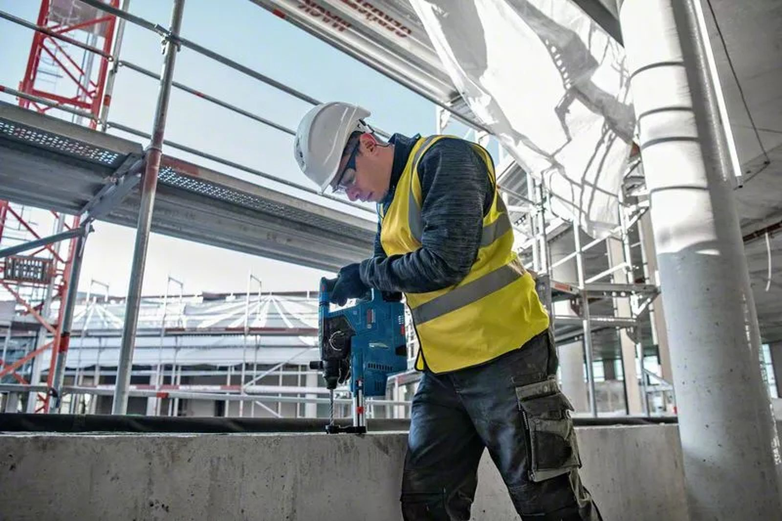 Ein Bauarbeiter bohrt in Beton. Er trägt einen Helm, eine Brille und eine gelbe Weste. Gerüste sind hinter ihm.