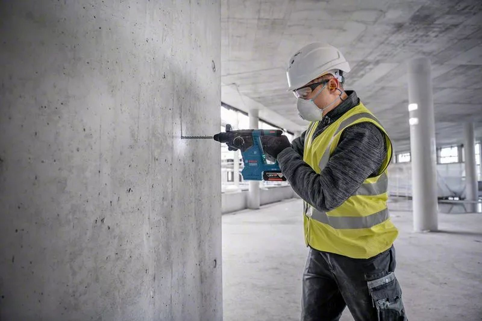 Ein Bauarbeiter bohrt in eine Betonmauer. Er trägt einen Helm, eine Schutzbrille, eine Maske und eine Warnweste.