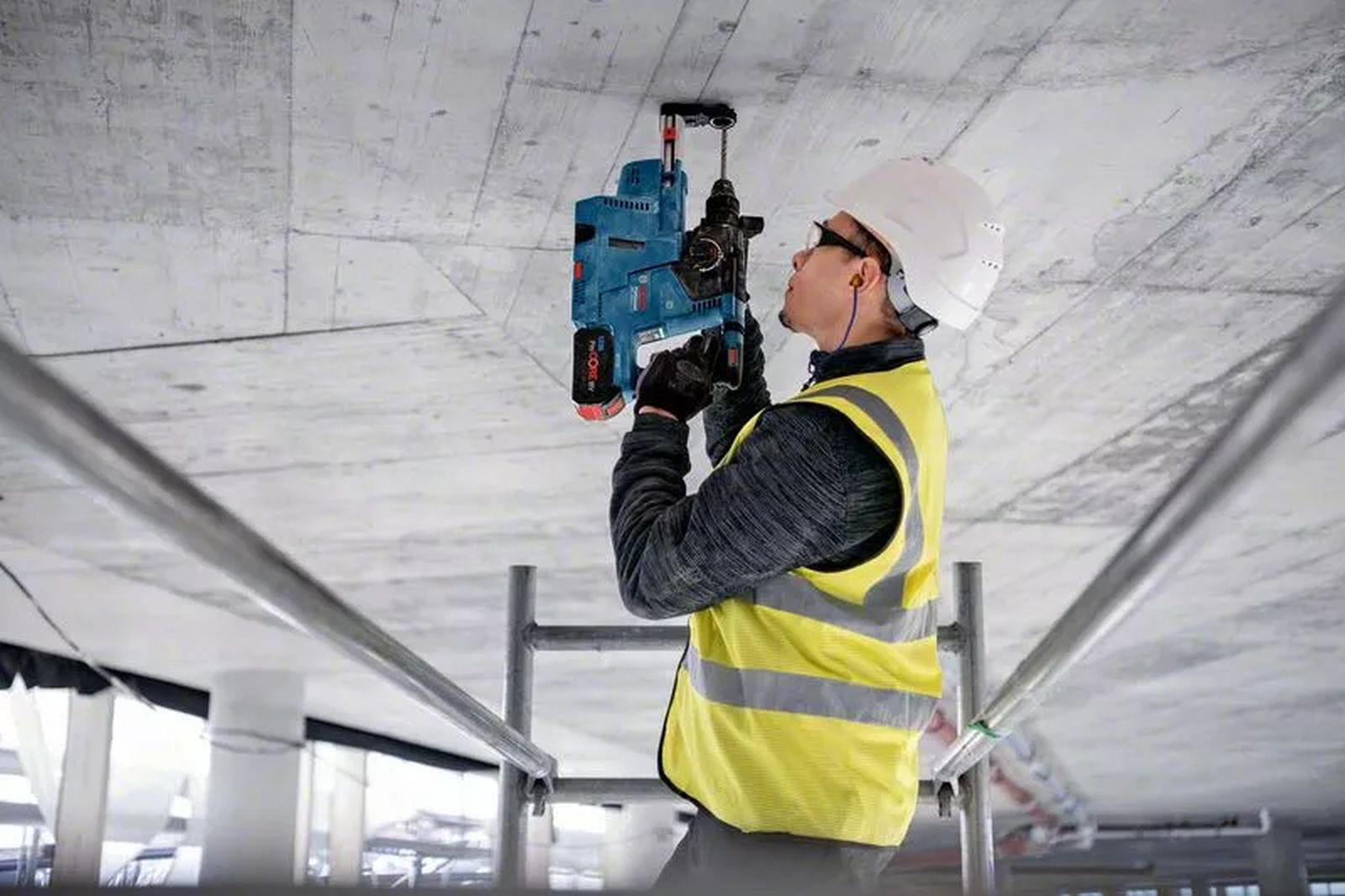 Ein Bauarbeiter bohrt mit einem blauen Elektrowerkzeug in eine Betondecke. Er trägt Helm und Sicherheitsweste.