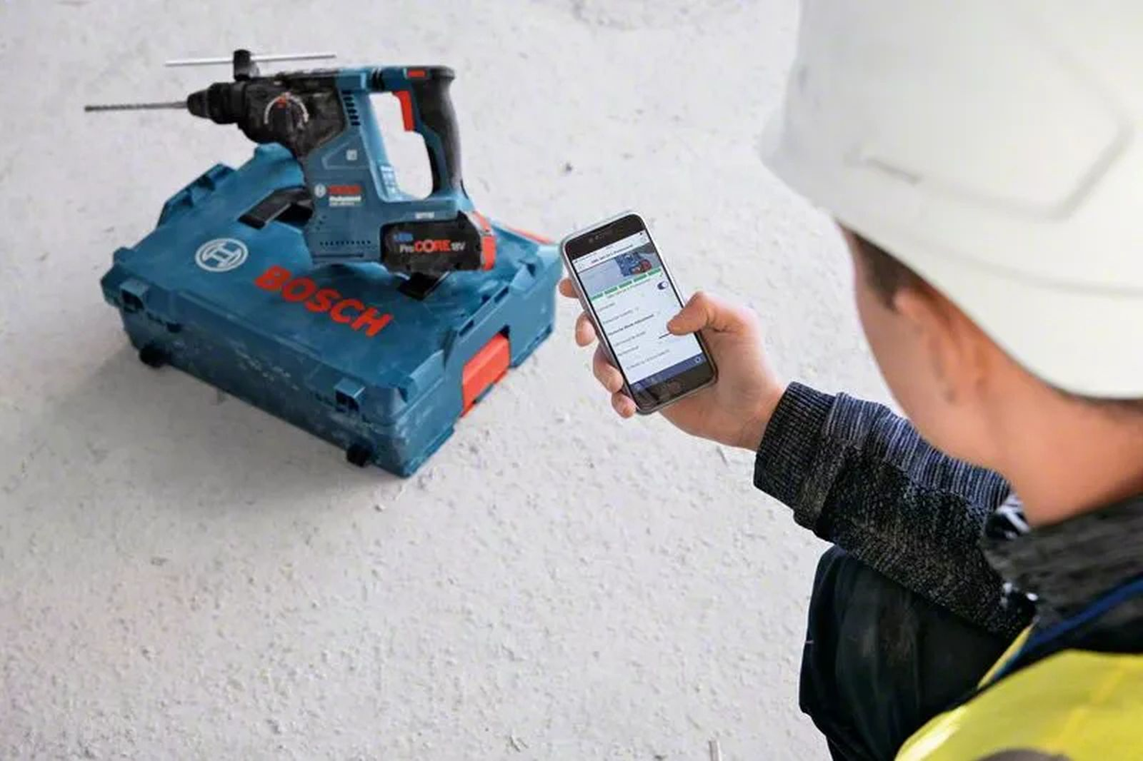 Eine Person mit Helm hält ein Telefon. Ein Bohrer liegt auf einem blauen Koffer. Die Szene spielt sich auf einer Baustelle ab.