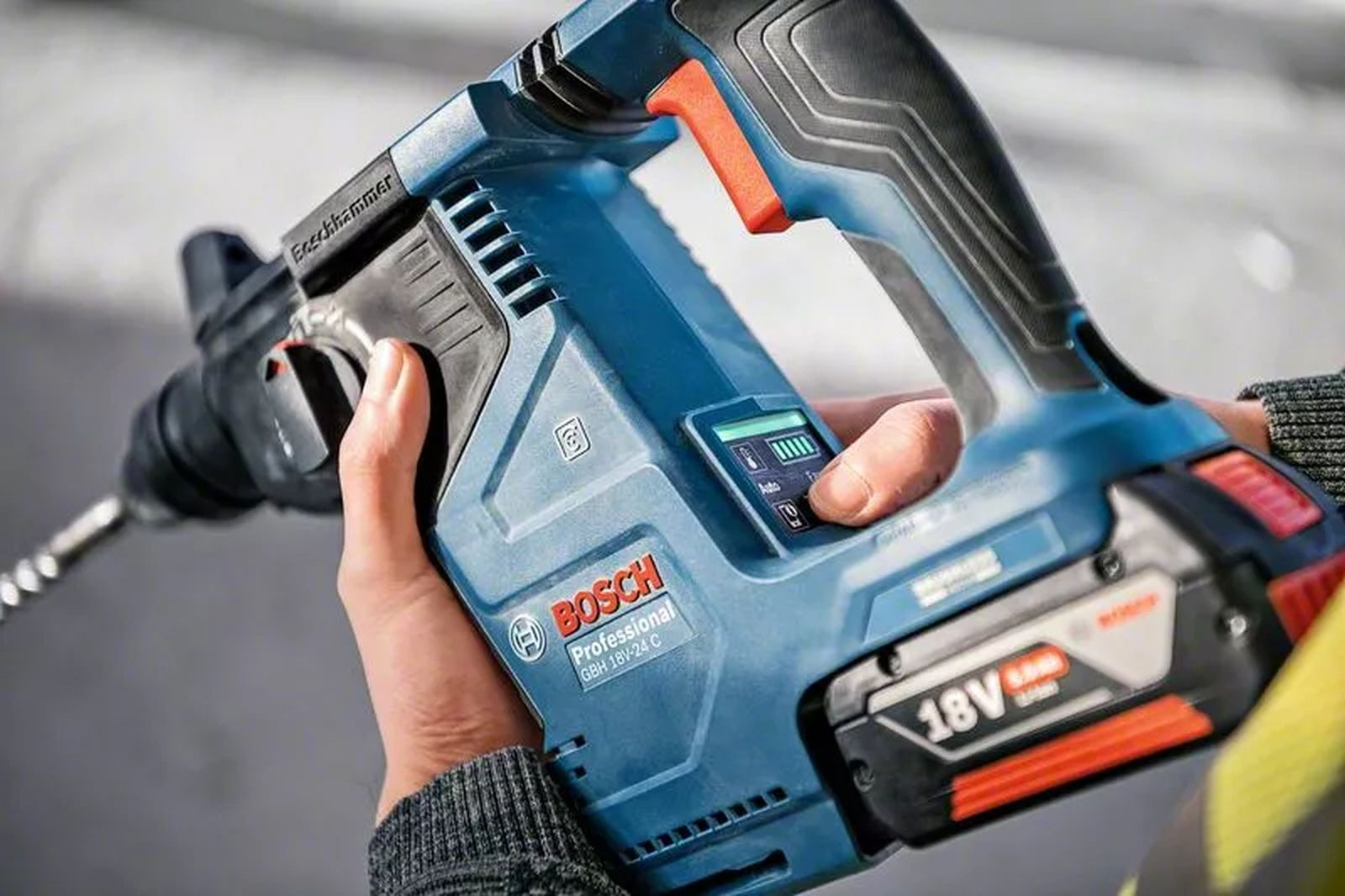 Eine blaue Bosch-Hammerbohrmaschine wird von Händen gehalten. Sie hat einen Akku und ein Bedienfeld. Der Bohrer ragt heraus.