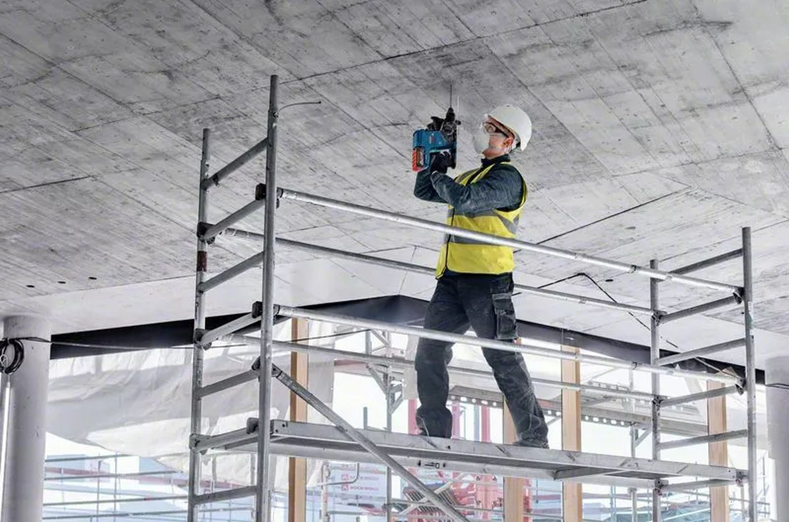 Ein Bauarbeiter bohrt von einem Gerüst aus in eine Betondecke. Er trägt Helm, Sicherheitsweste und Schutzbrille.
