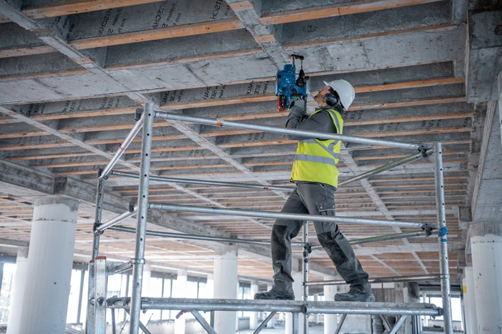 Robotnik budowlany używa wiertarki do betonowego sufitu, stojąc na rusztowaniu. Nosi kask i kamizelkę odblaskową.