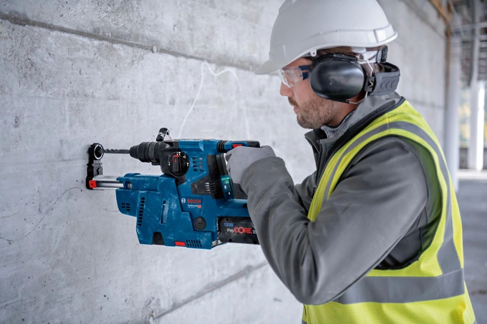 Robotnik budowlany wierci w betonowej ścianie niebieską wiertarką. Nosi okulary ochronne, ochronniki słuchu i kamizelkę.