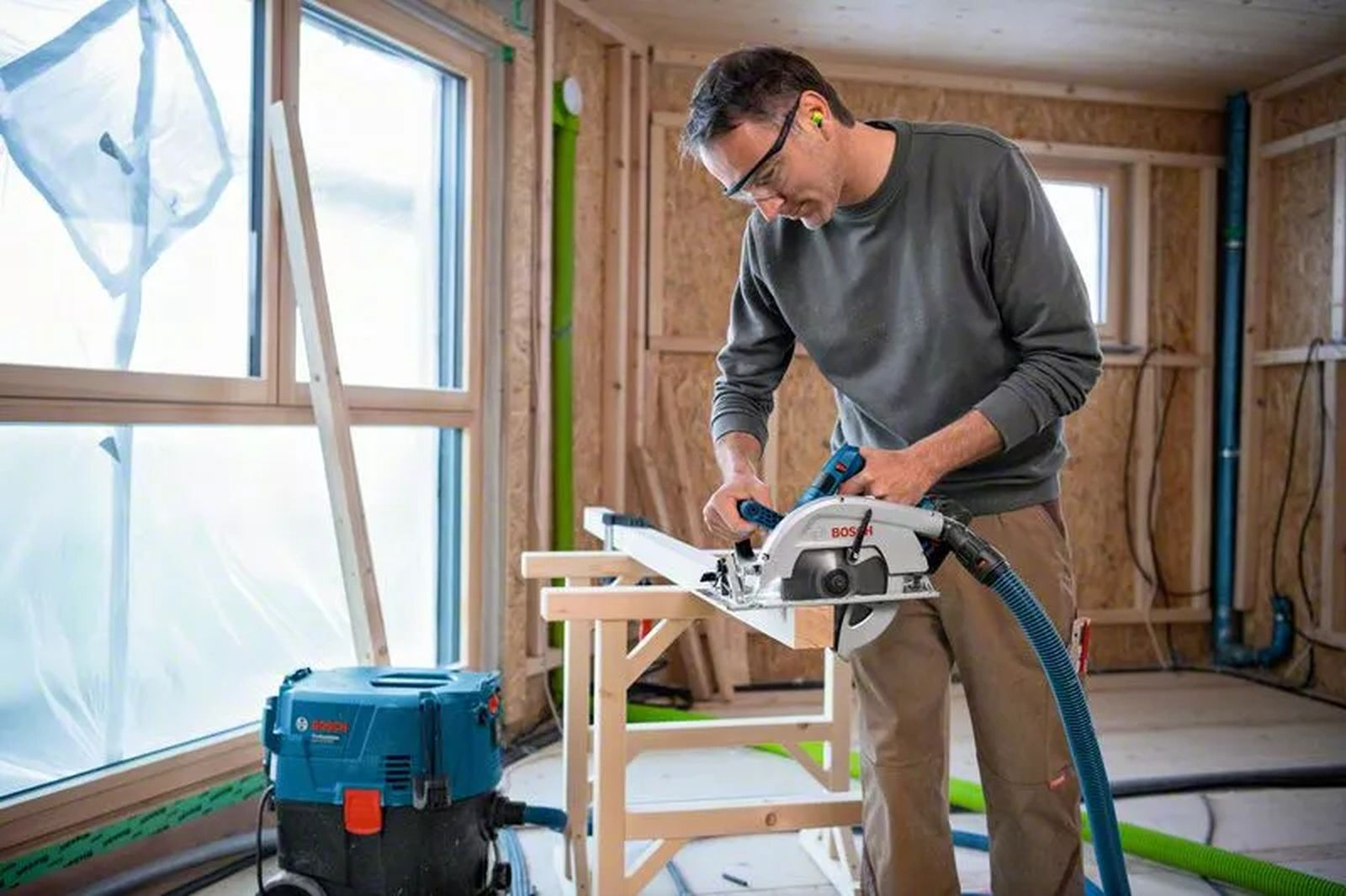 Mężczyzna używa piły tarczowej do drewna. Nosi okulary i zatyczki do uszu. Odkurzacz jest w pobliżu.