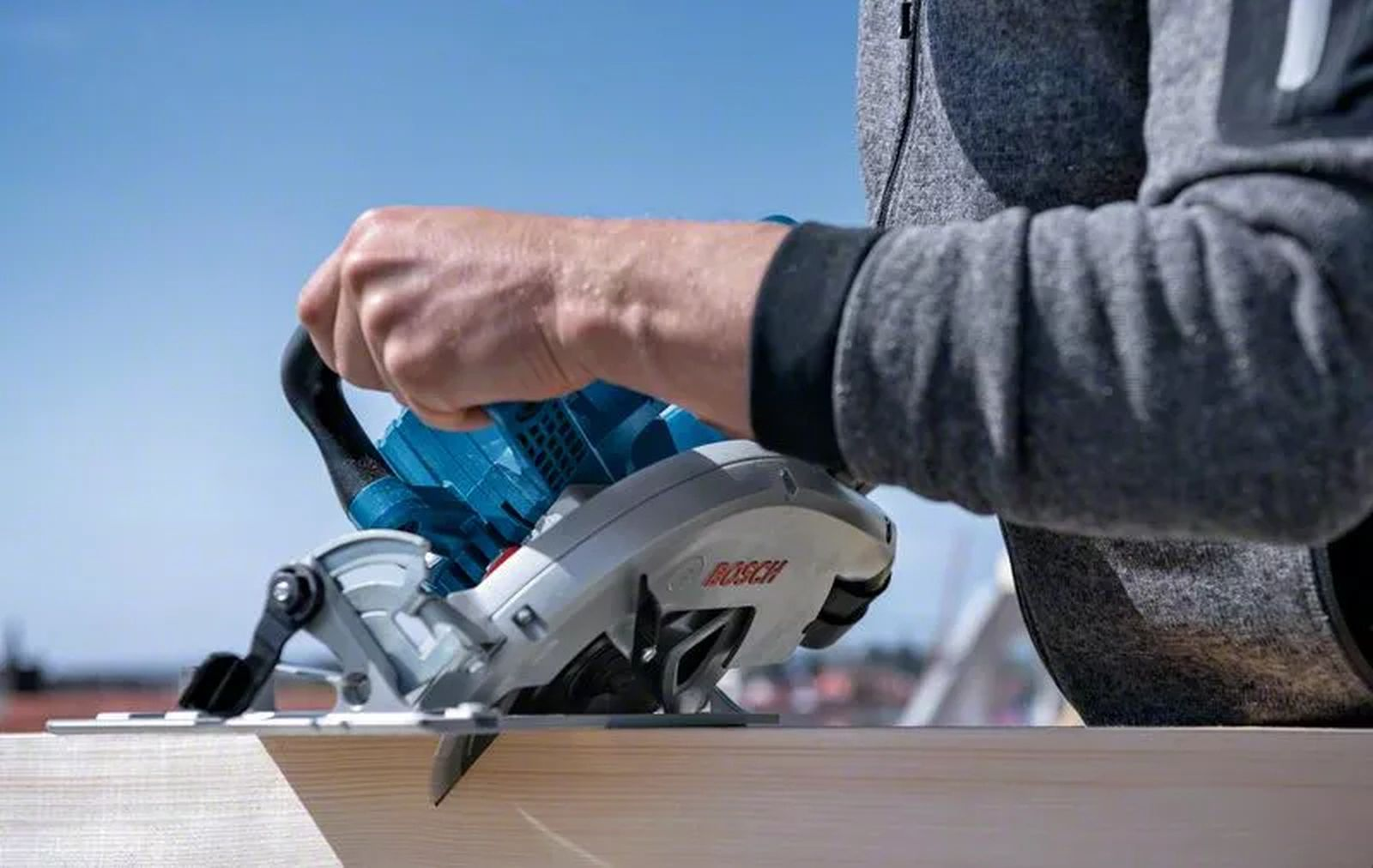 Osoba używa niebiesko-srebrnej piły tarczowej do cięcia drewnianej deski na tle błękitnego nieba.