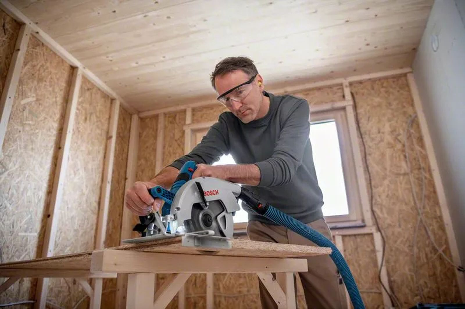 Mężczyzna używa piły tarczowej do cięcia drewna. Ma okulary ochronne i ochronniki słuchu. Scena ma miejsce w budowie.