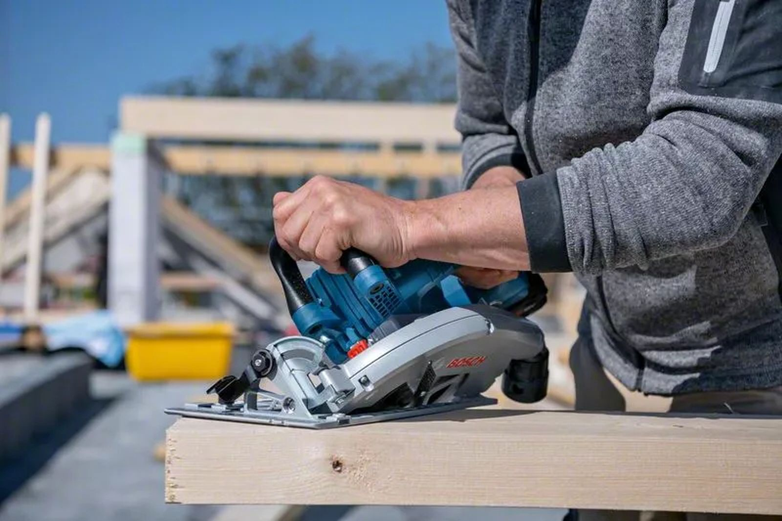 Osoba używa niebiesko-srebrnej piły tarczowej do cięcia drewnianej deski. W tle widać budowę.