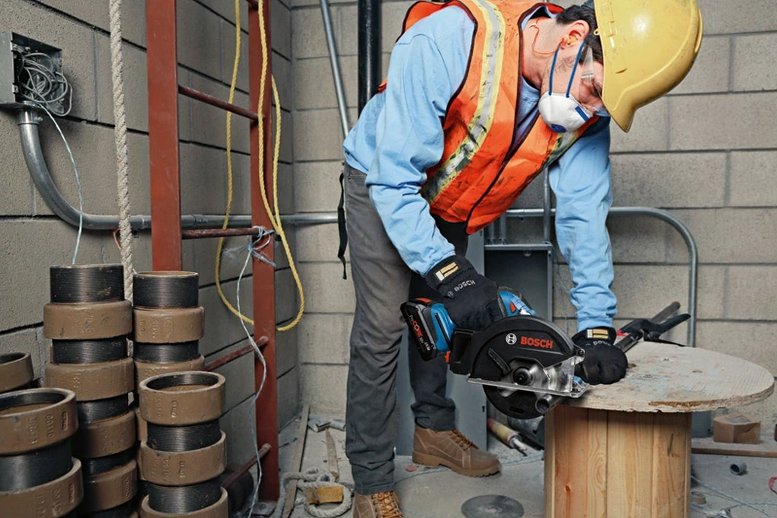 Robotnik budowlany używa piły tarczowej. Nosi sprzęt ochronny. Po lewej stronie stos metalowych pierścieni. Po prawej drewniany stół szpulowy.
