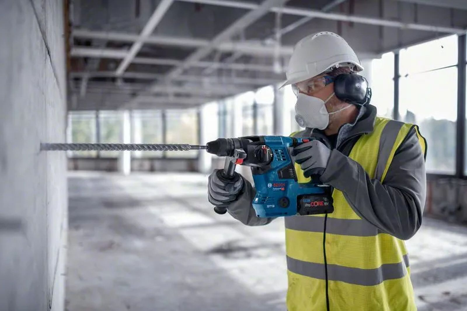 Robotnik budowlany wierci w betonowej ścianie niebieską wiertarką, nosząc odzież ochronną, w budynku.