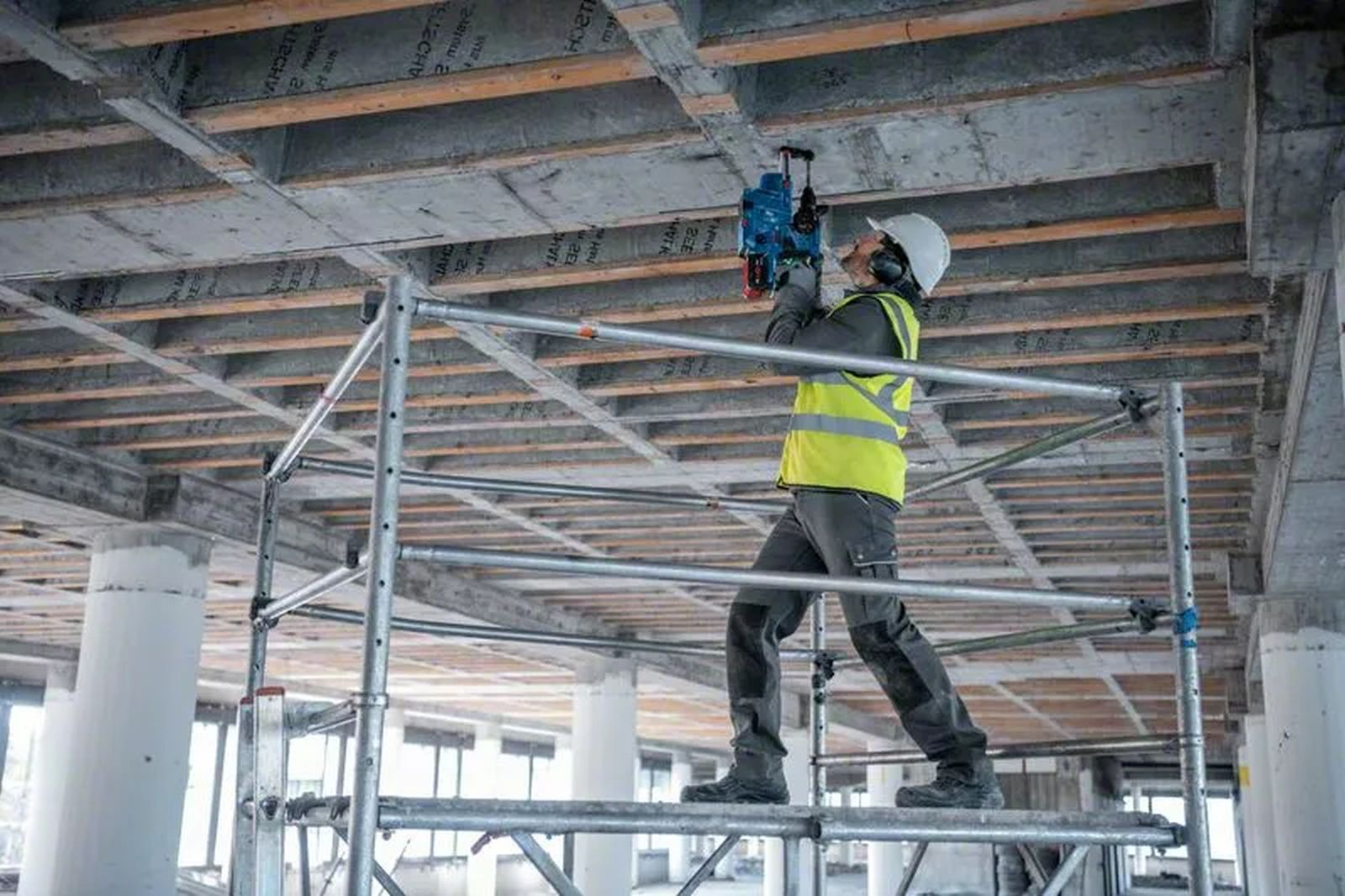 Robotnik budowlany wierci w betonowym suficie. Ma na sobie kask, ochronniki słuchu i żółtą kamizelkę odblaskową, stojąc na rusztowaniu.