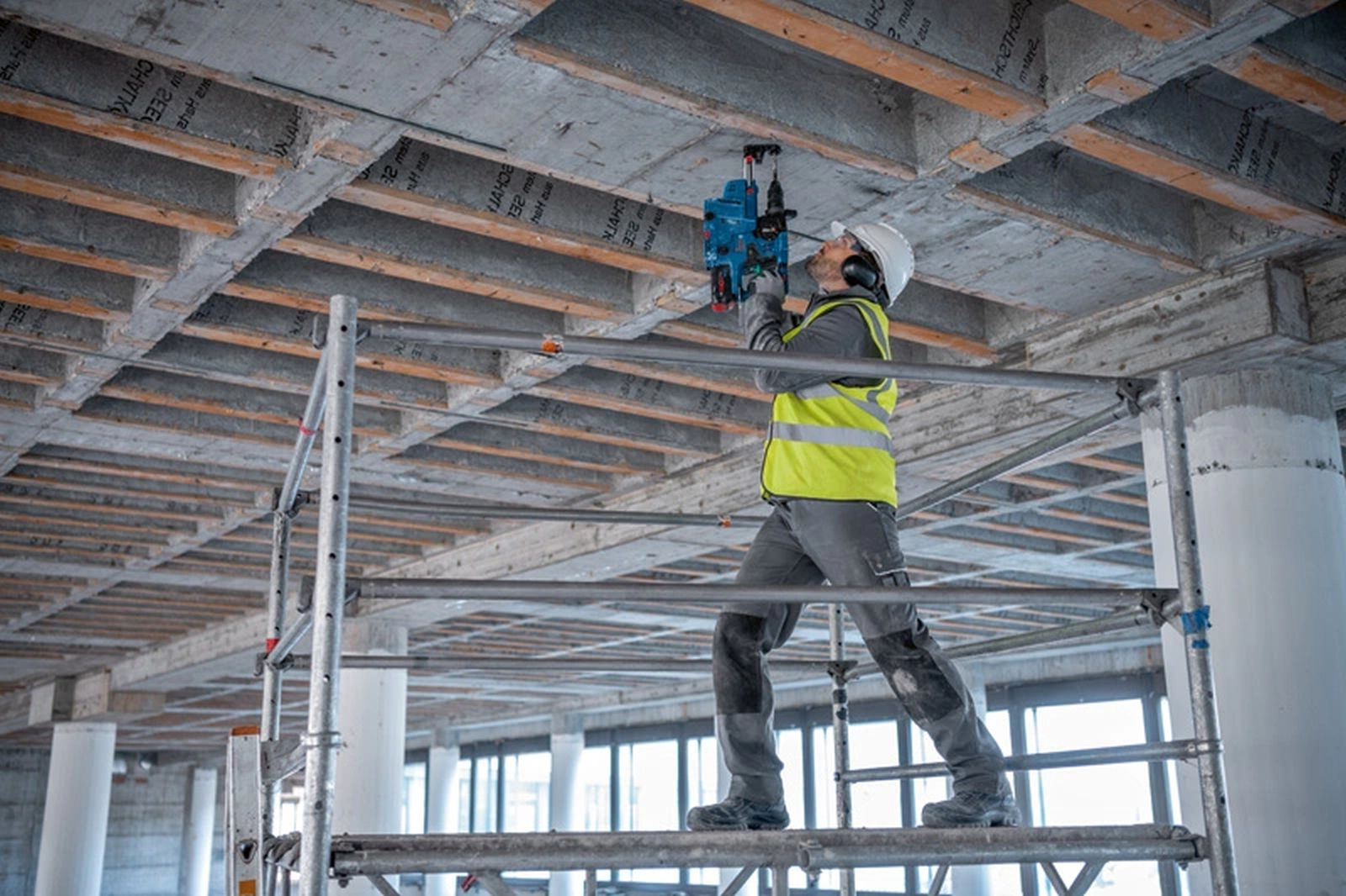 Robotnik budowlany wierci w betonowym suficie. Ma na sobie kamizelkę, kask i ochronę słuchu, stojąc na rusztowaniu.