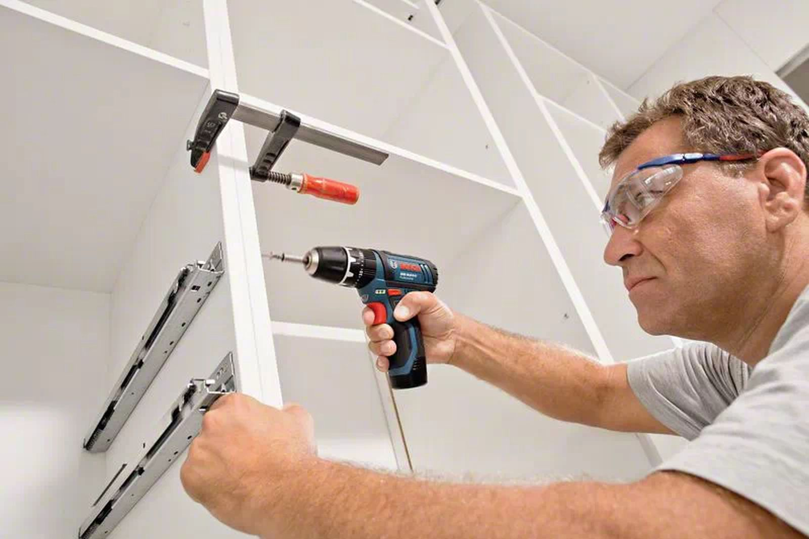Un homme utilise une perceuse sur une armoire. Il porte des lunettes de sécurité. Une pince maintient l'armoire. Le fond est blanc.