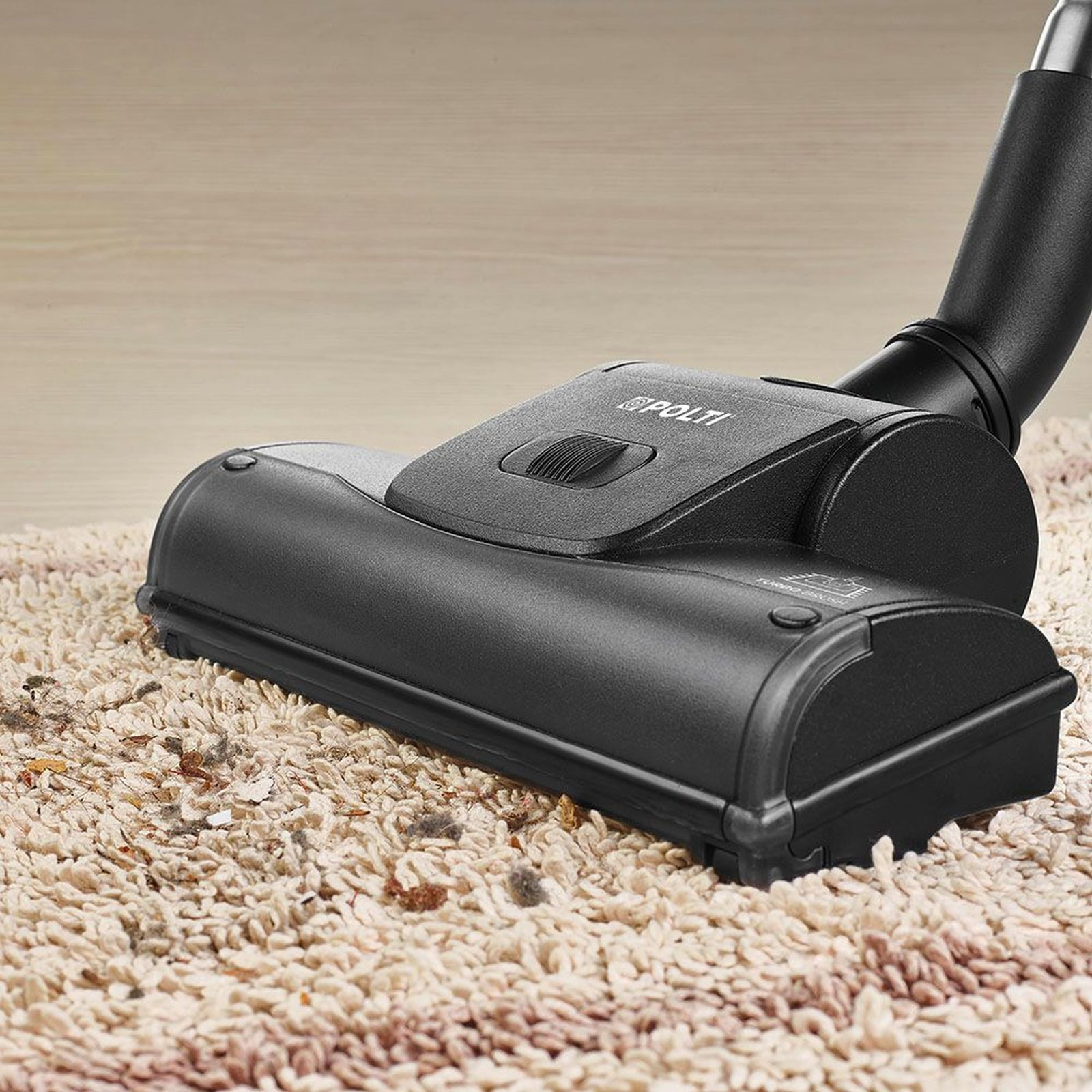 Czarny odkurzacz Polti na beżowym dywanie, widoczny kurz i zanieczyszczenia. Tło to drewniana podłoga.