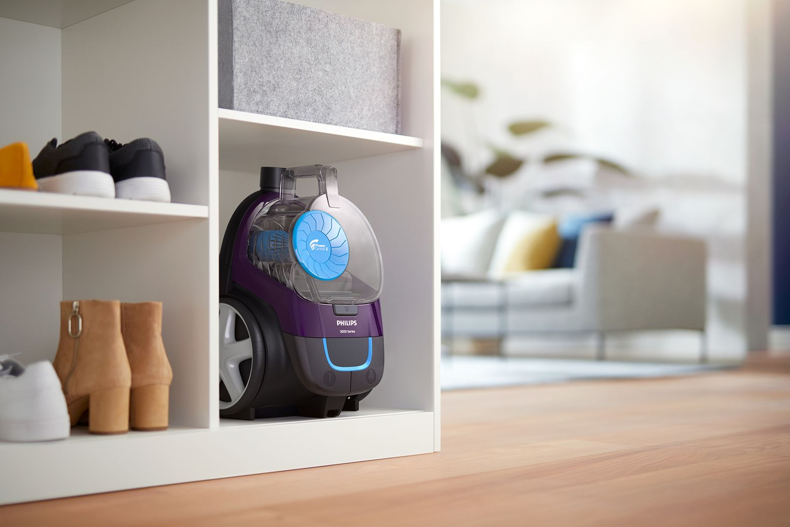 Fioletowy odkurzacz Philips stoi na białej półce. Na półce są również buty. W tle znajduje się salon.