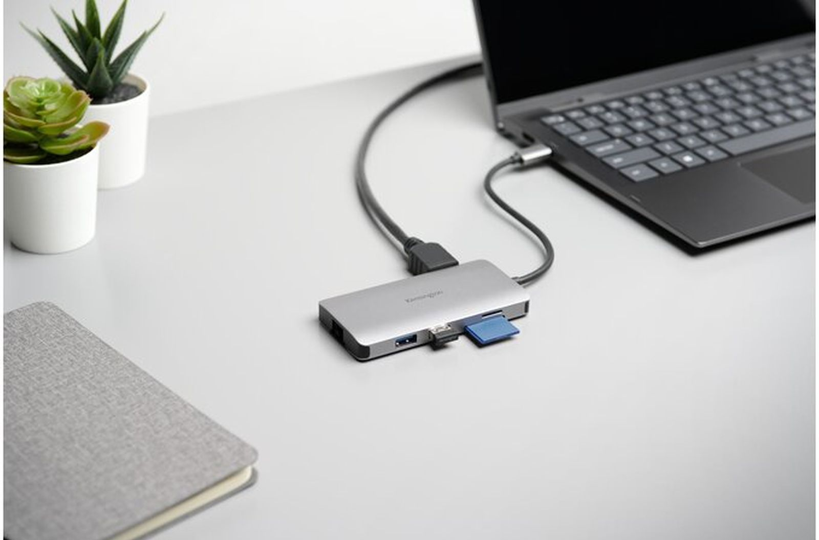 Un laptop collegato a un hub argentato con una scheda SD blu. Piante e un taccuino sulla scrivania bianca.