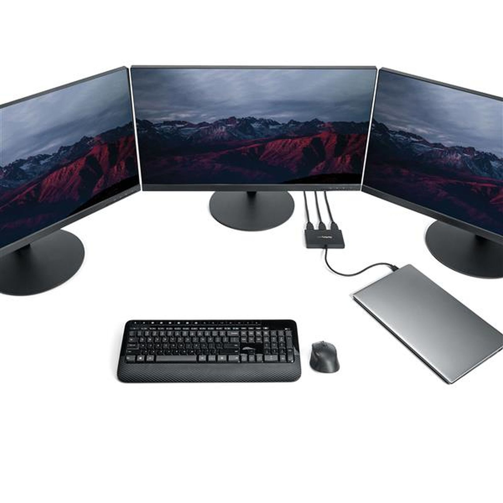 Tres monitores muestran una escena de montaña. Teclado, ratón y portátil sobre una superficie blanca.