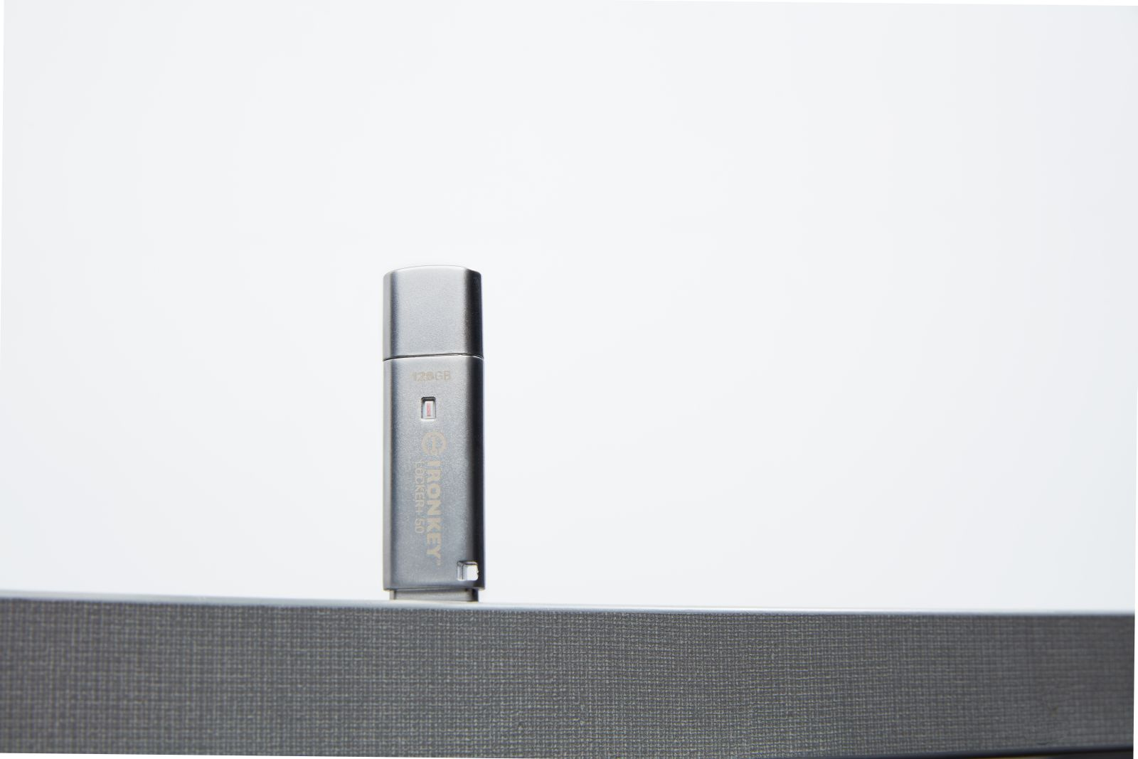 Srebrny pendrive stoi pionowo na szarej powierzchni na białym tle. Dysk ma napis: IronKey Locker+ 50, 128 GB.