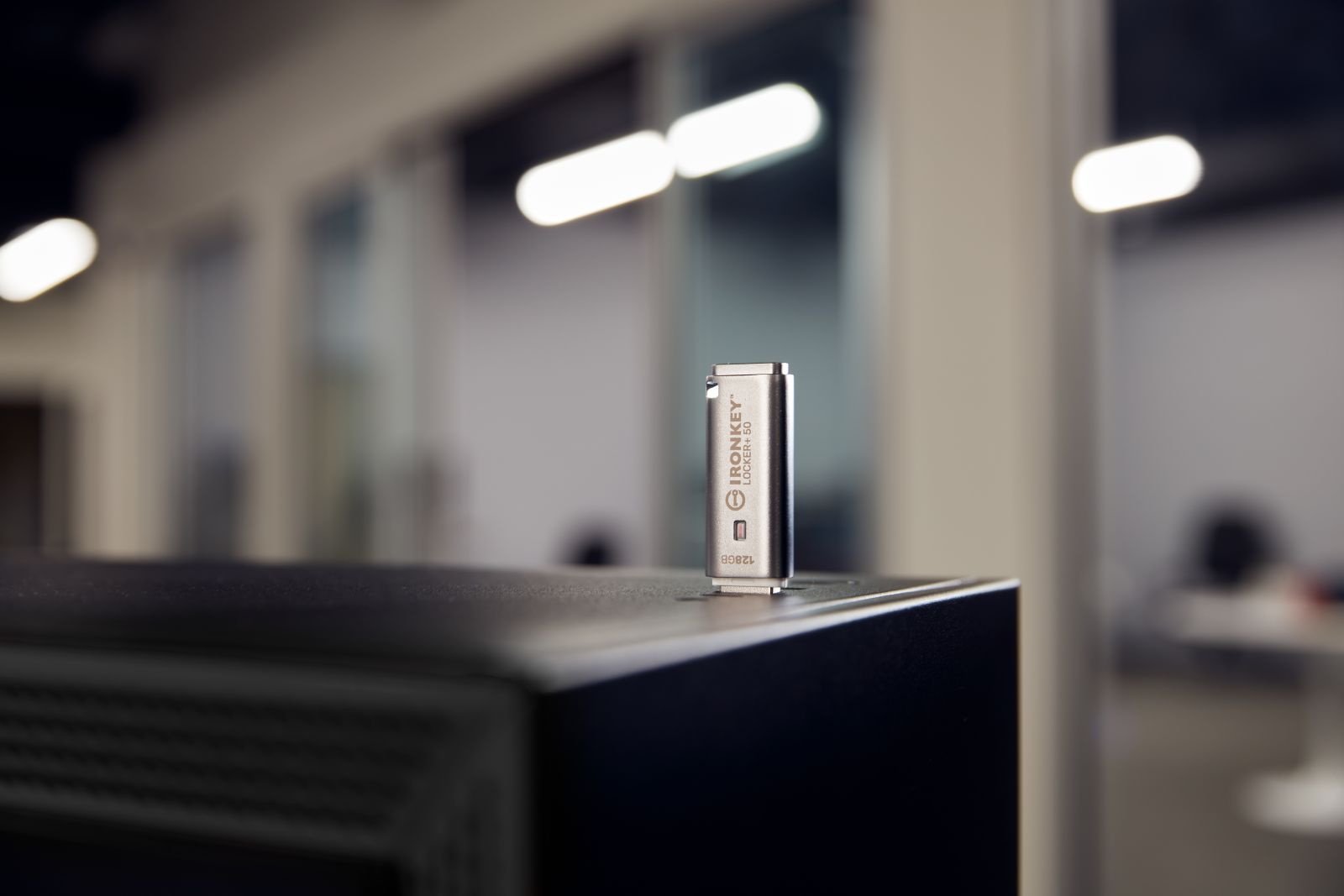 Srebrny pendrive IronKey stoi na czarnej powierzchni. Tło przedstawia biuro z rozmytymi światłami i ścianami.