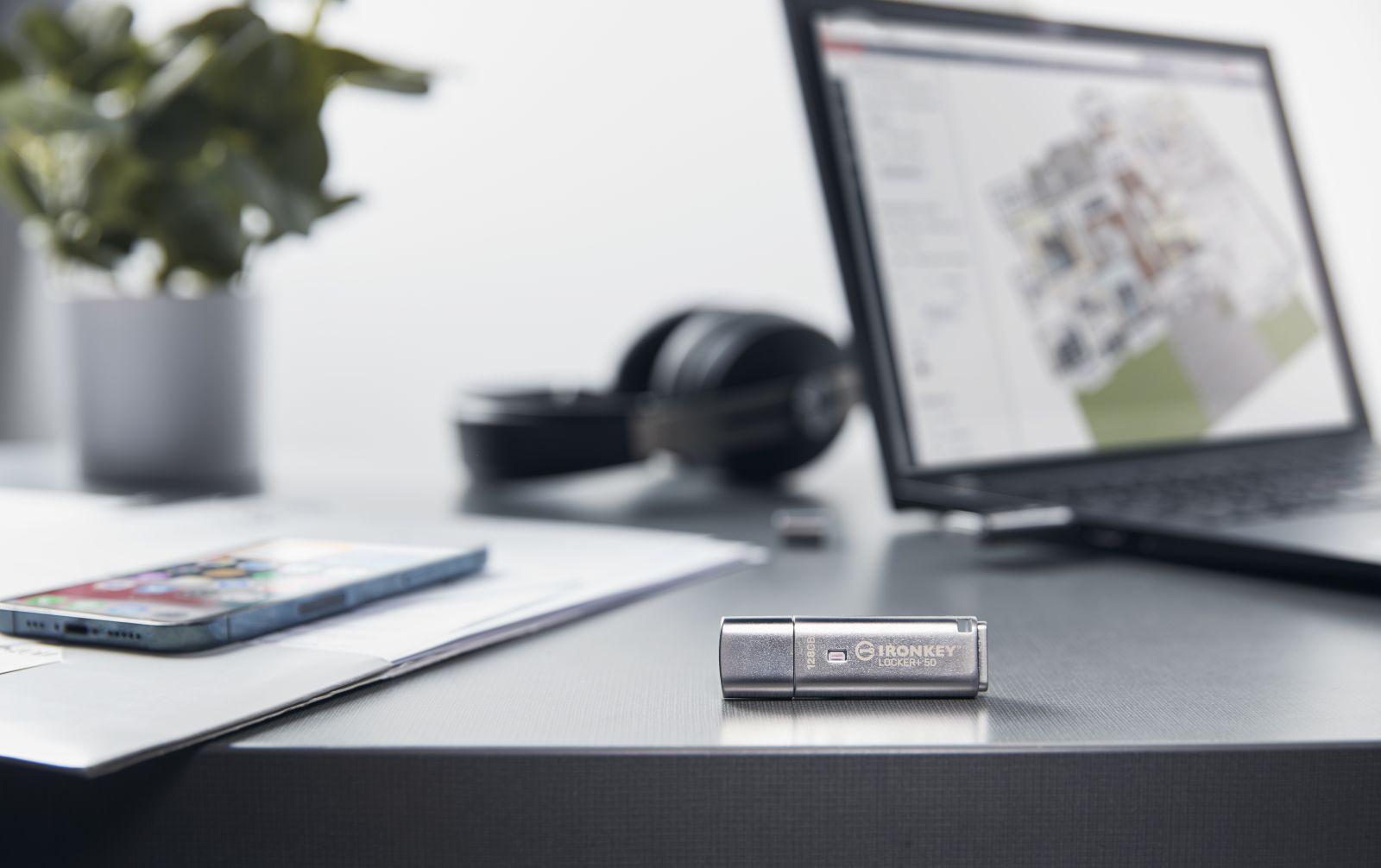 Biurko z laptopem, słuchawkami, telefonem i pendrivem. Laptop wyświetla plan. Roślina w tle.