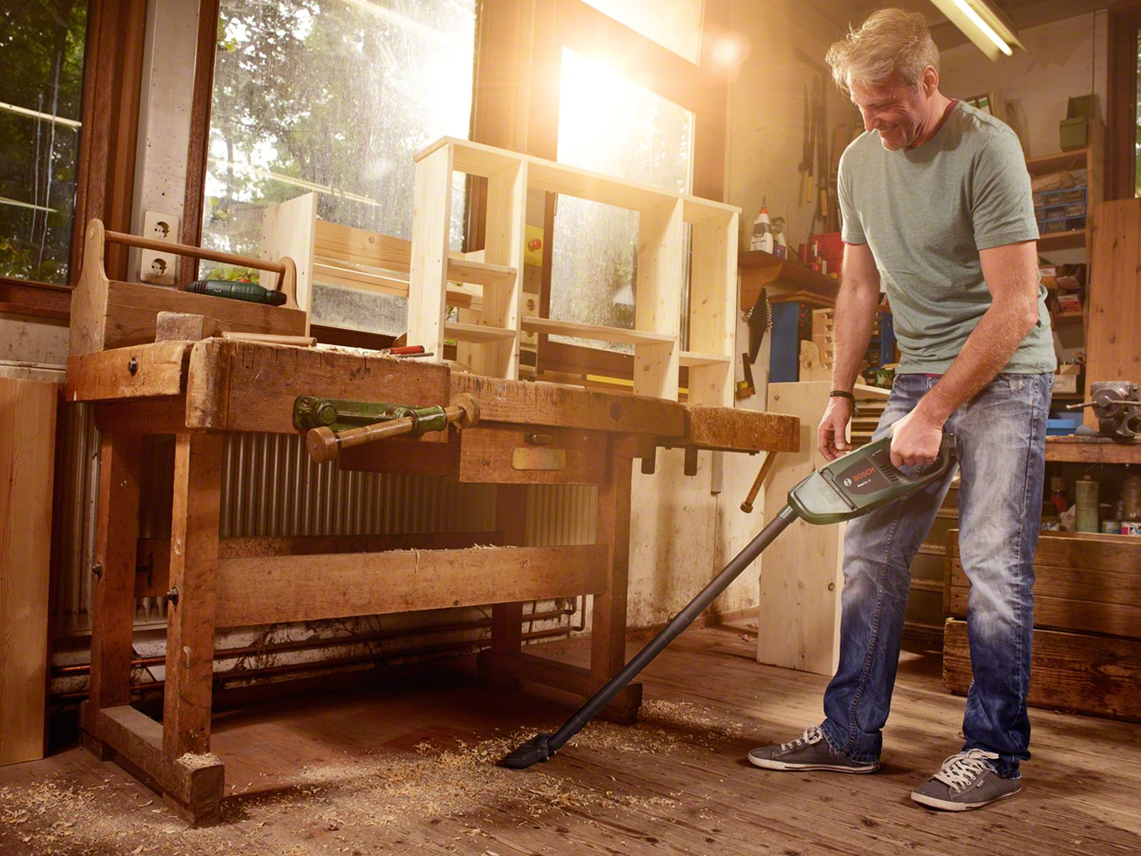 Un hombre aspira virutas de madera en un taller. Un banco de trabajo, estanterías y herramientas están al fondo. La luz solar entra.