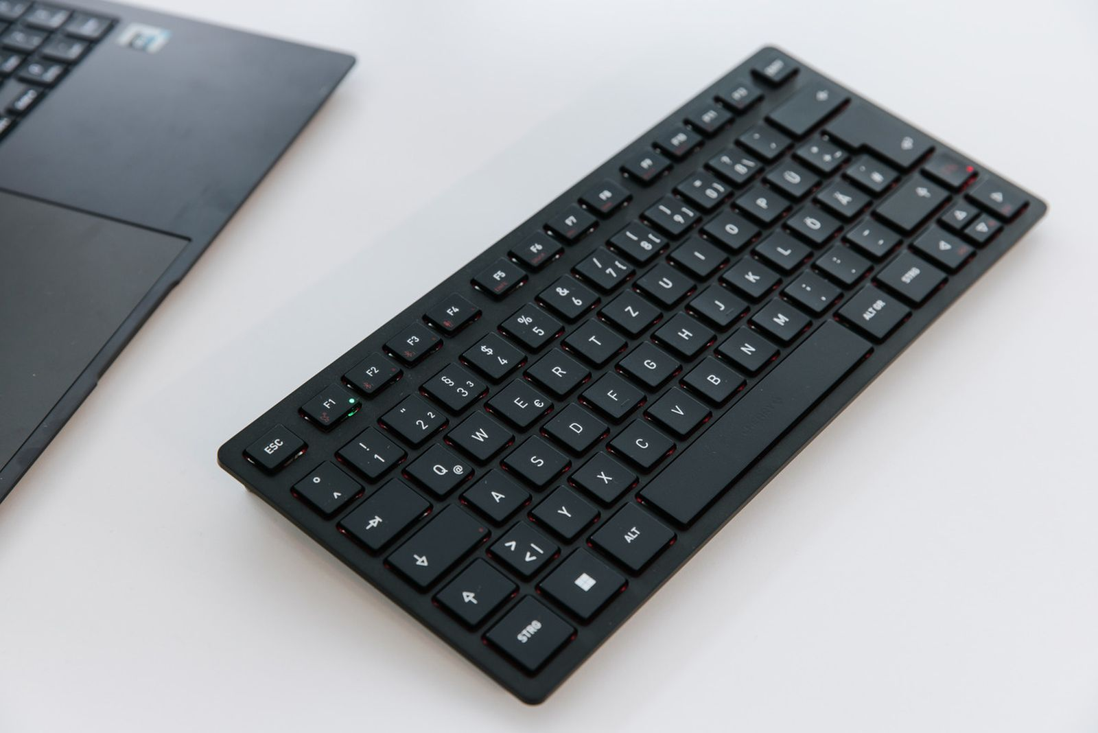 Een zwart toetsenbord met witte toetsen en rode accenten ligt op een wit oppervlak naast een zwarte laptop.