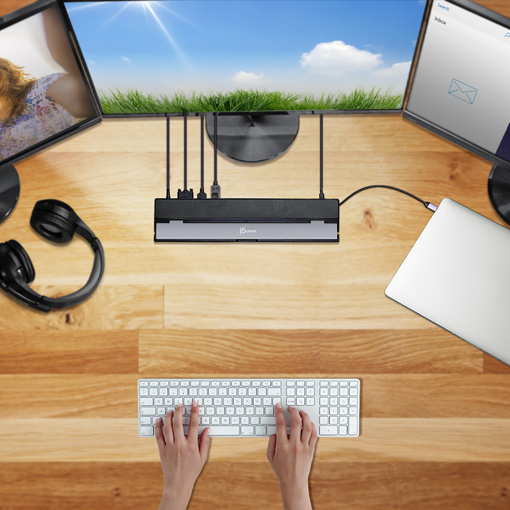 Ein Schreibtisch mit drei Monitoren, Laptop, Tastatur und Kopfhörern. Eine Person tippt.