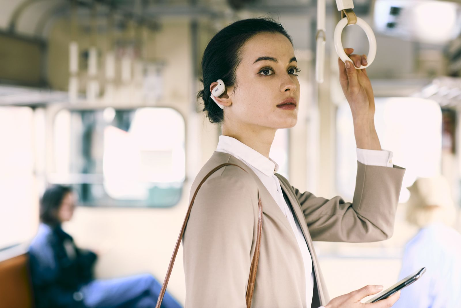 Kobieta w pociągu trzyma uchwyt, ma na sobie słuchawki i marynarkę. Trzyma telefon. Inni są w tle.