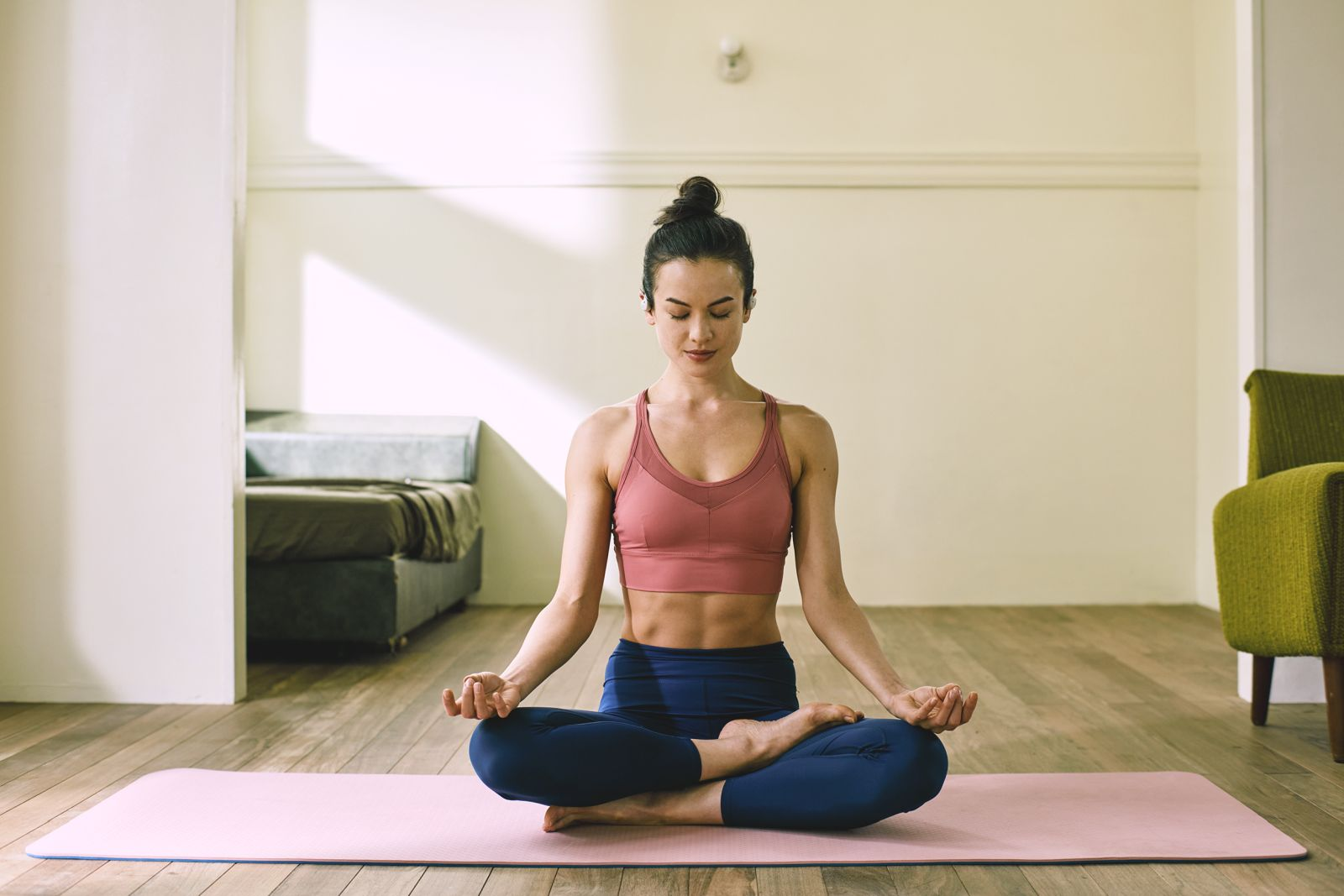 Kobieta w pozycji jogi, pozycja lotosu, na różowej macie. Ubrana w różowy top i niebieskie spodnie. Drewniana podłoga, zielone krzesło.