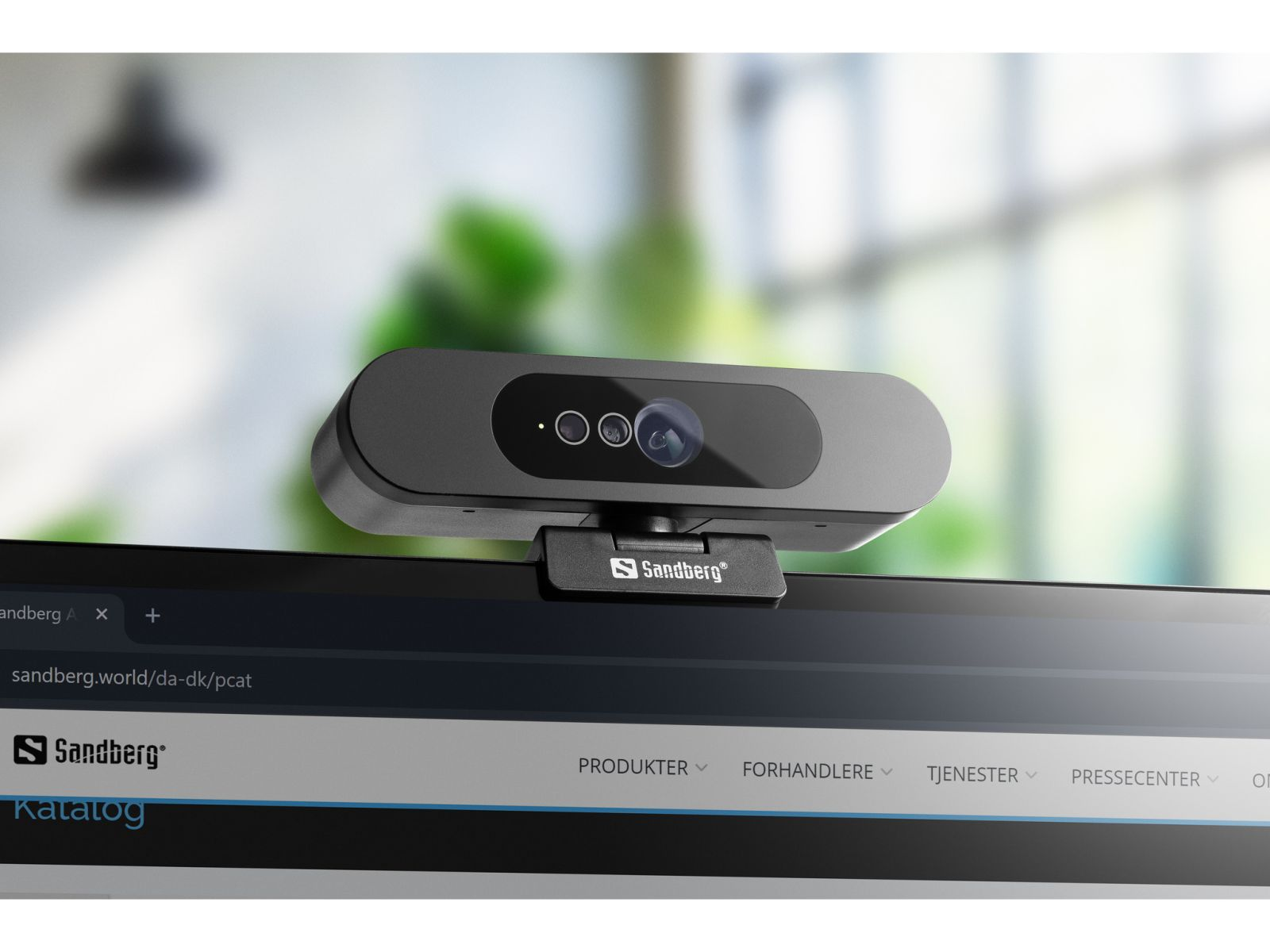 Kamera internetowa Sandberg znajduje się na monitorze. Kamera jest czarna z trzema soczewkami. Tło to rozmyte okno.