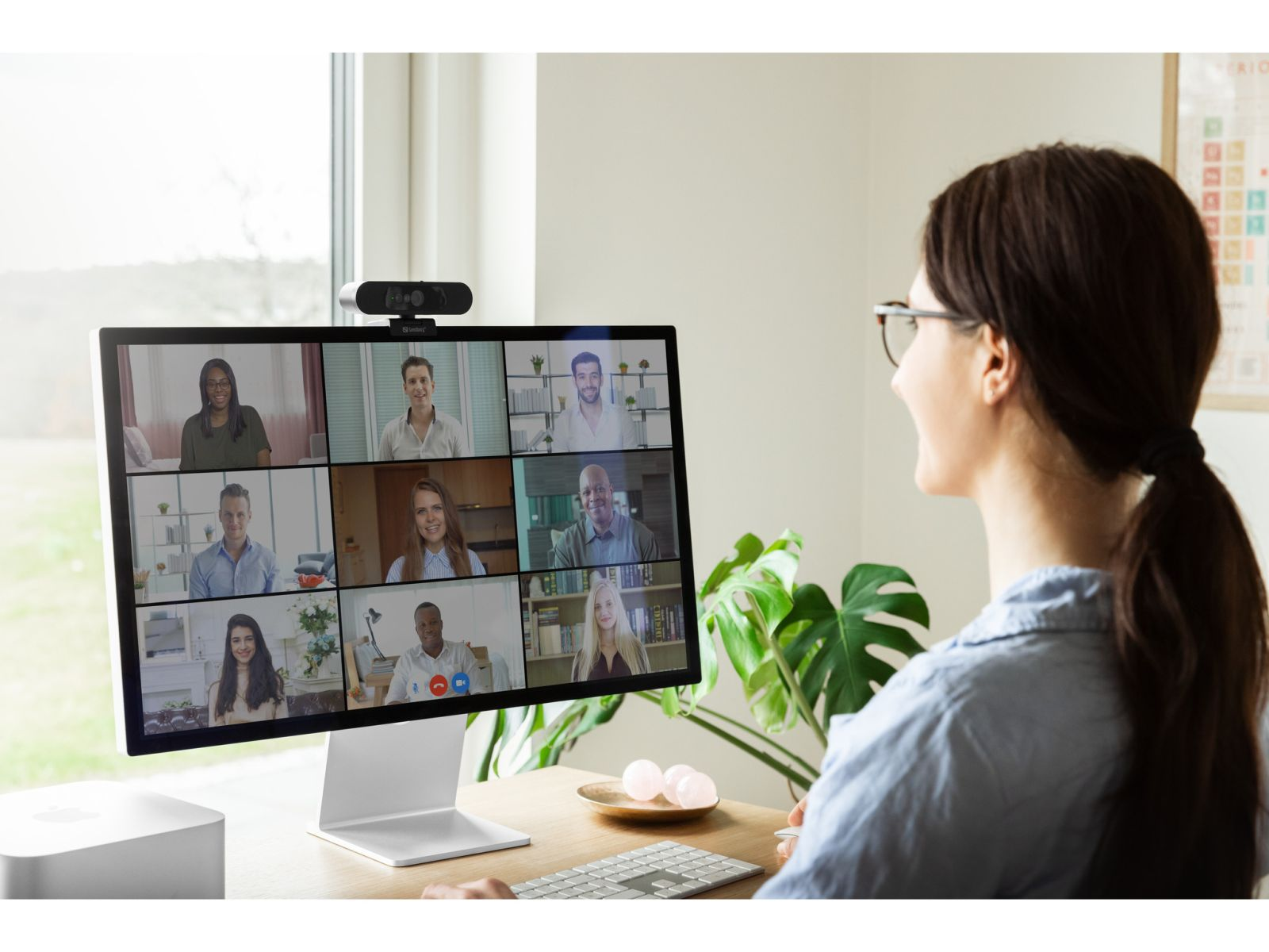 Kobieta patrzy na ekran komputera, na którym wyświetlana jest wideokonferencja. Dziewięć osób bierze udział w rozmowie wideo. Na górze jest kamera internetowa.